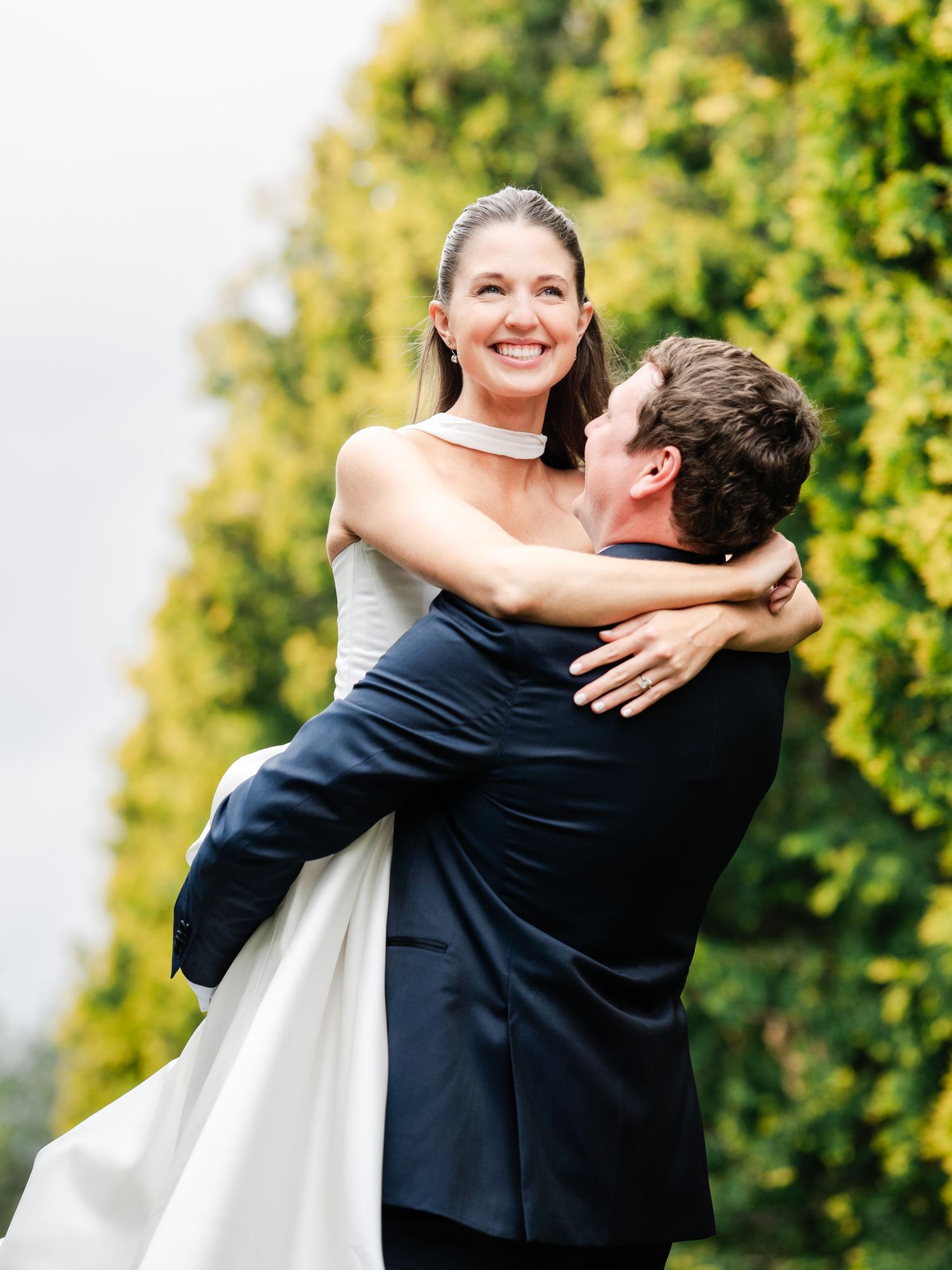 Groom lifting smiling bride in white wedding gown outdoors with lush green trees in background.