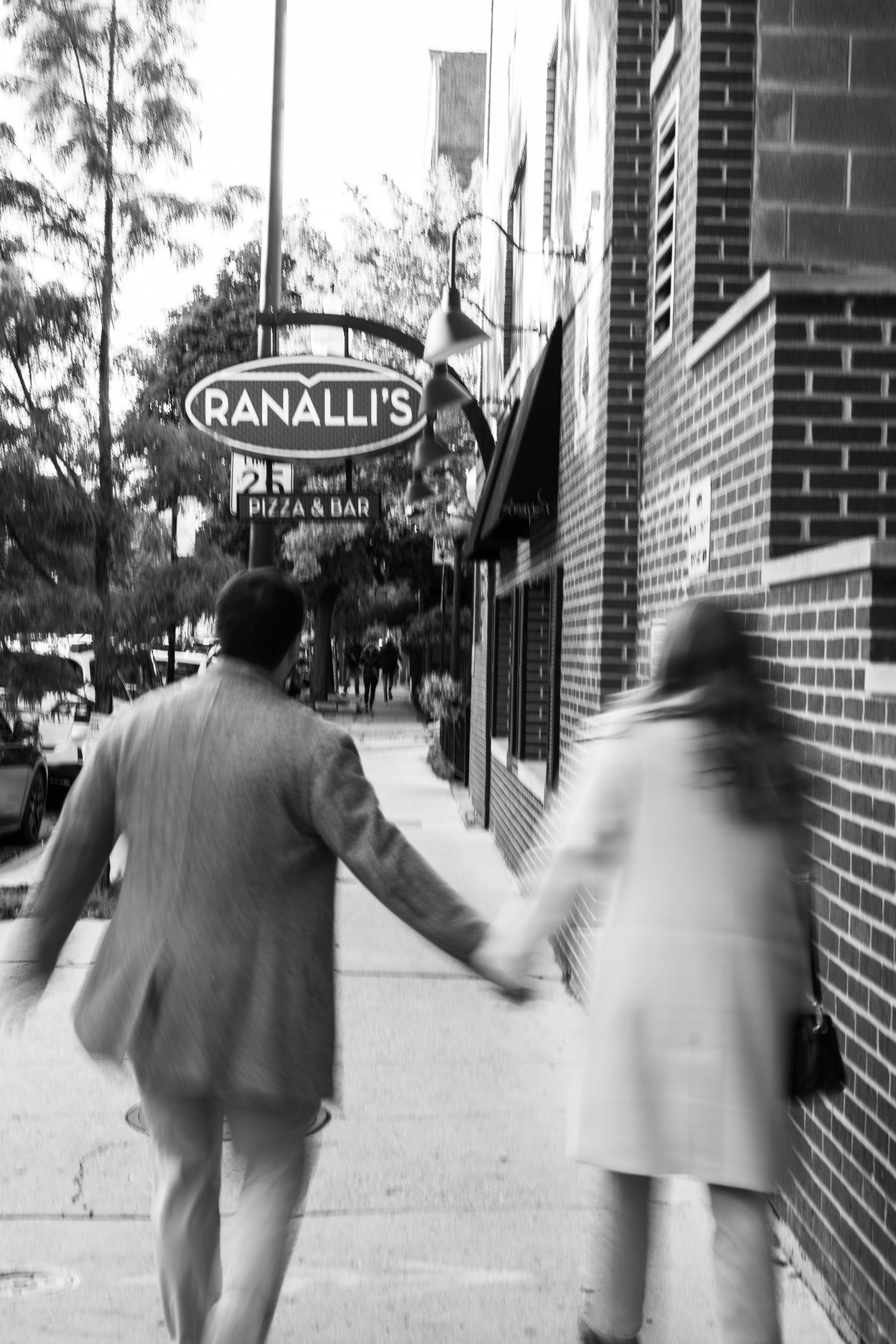 Couple walking hand in hand past Ranalli's Pizza and Bar sign in black and white street photo.