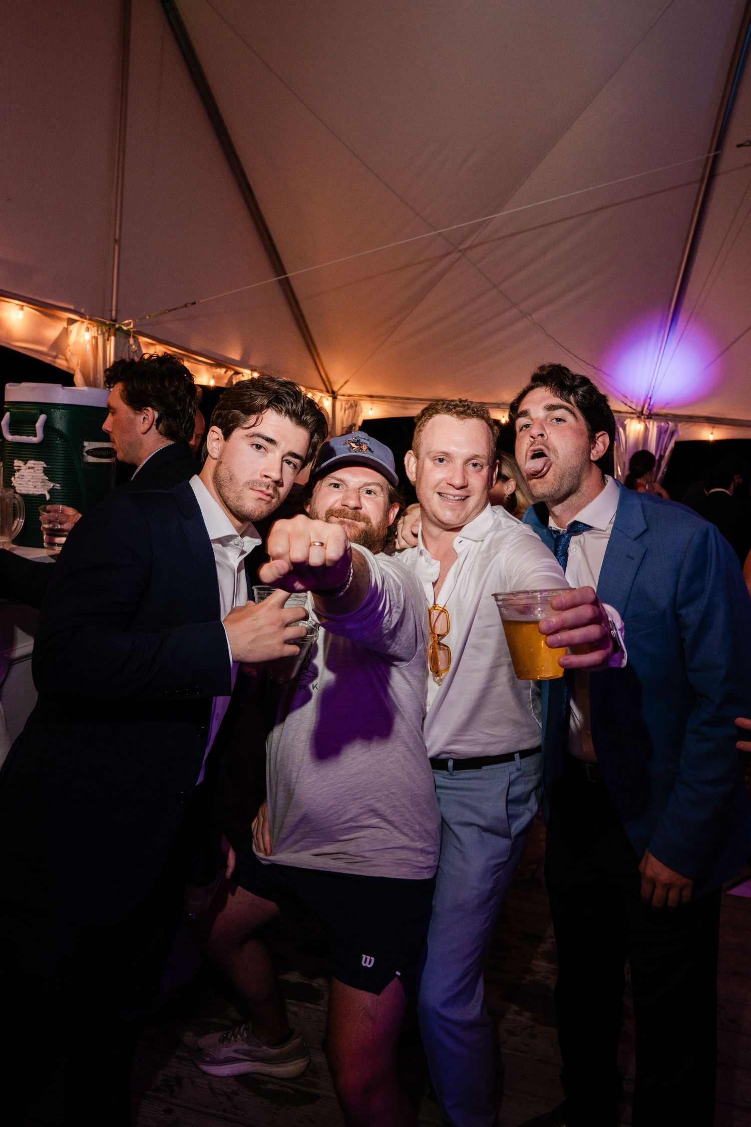 Four young men pose and celebrate together at a lively wedding reception under a tent.