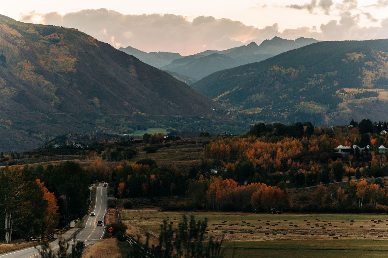 Scenic autumn valley landscape with winding road, golden trees, and mountain range under dramatic sunset sky.