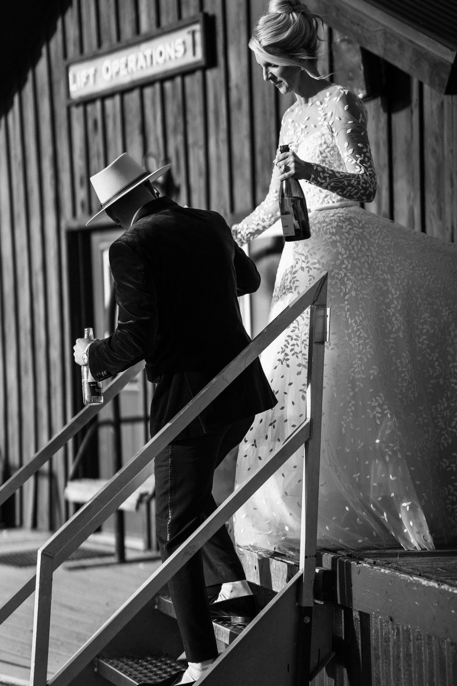 Black and white photo of bride and groom walking down stairs holding wine bottles at rustic venue.