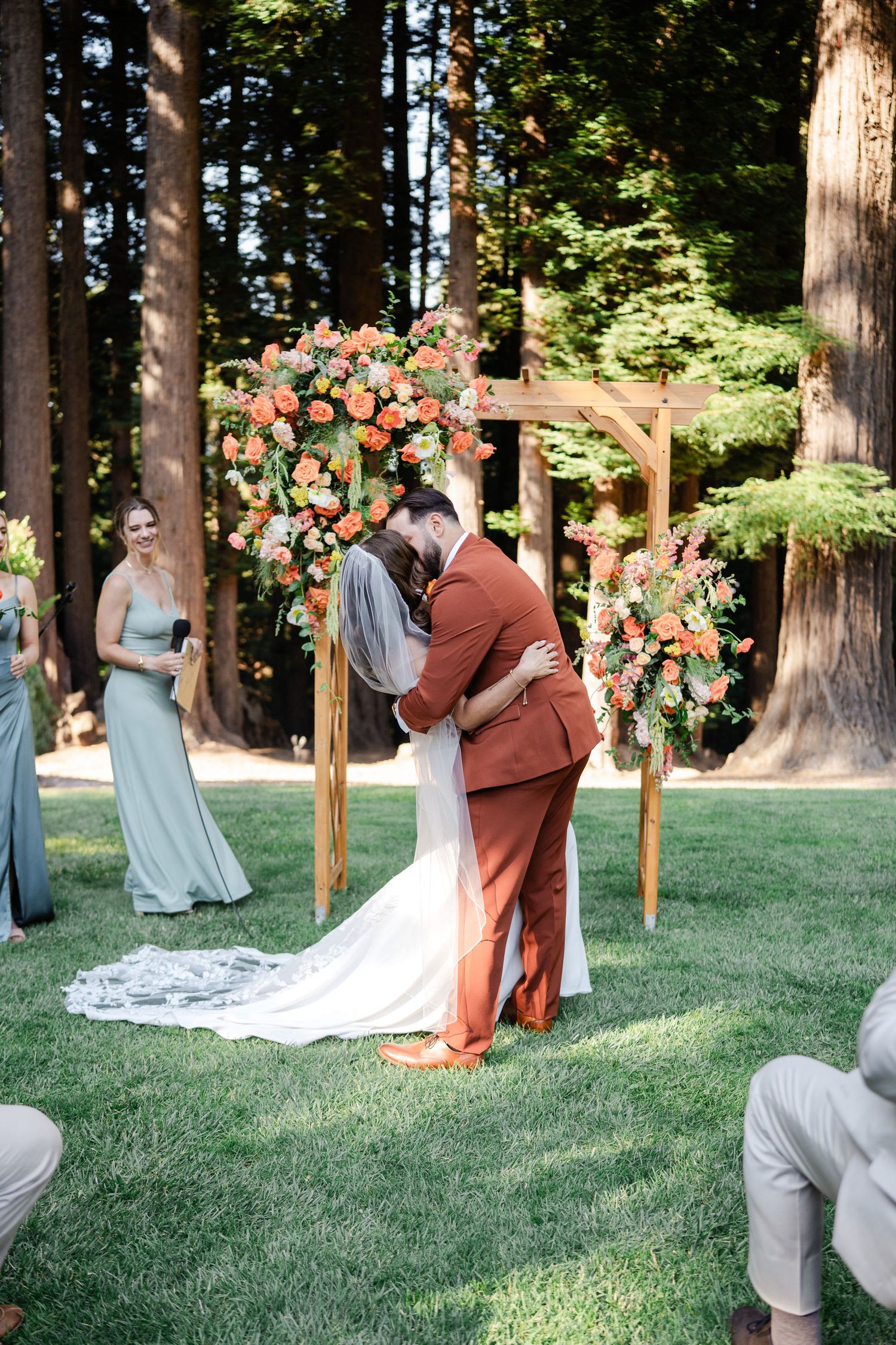 Bride and groom sharing first kiss at wooden floral arch surrounded by redwood trees.