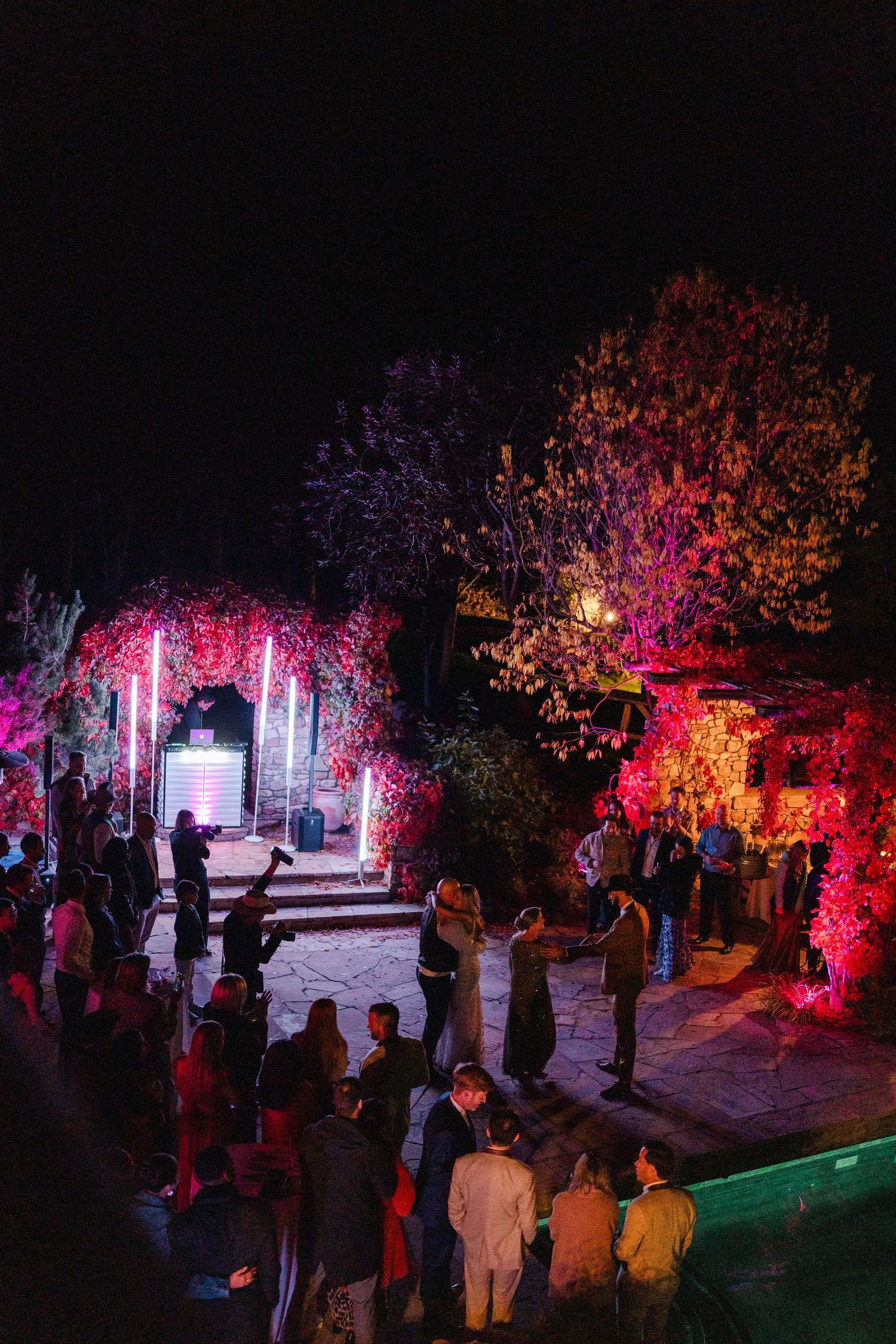 Outdoor evening wedding reception with colorful pink and red lighting illuminating trees and guests mingling on stone patio.