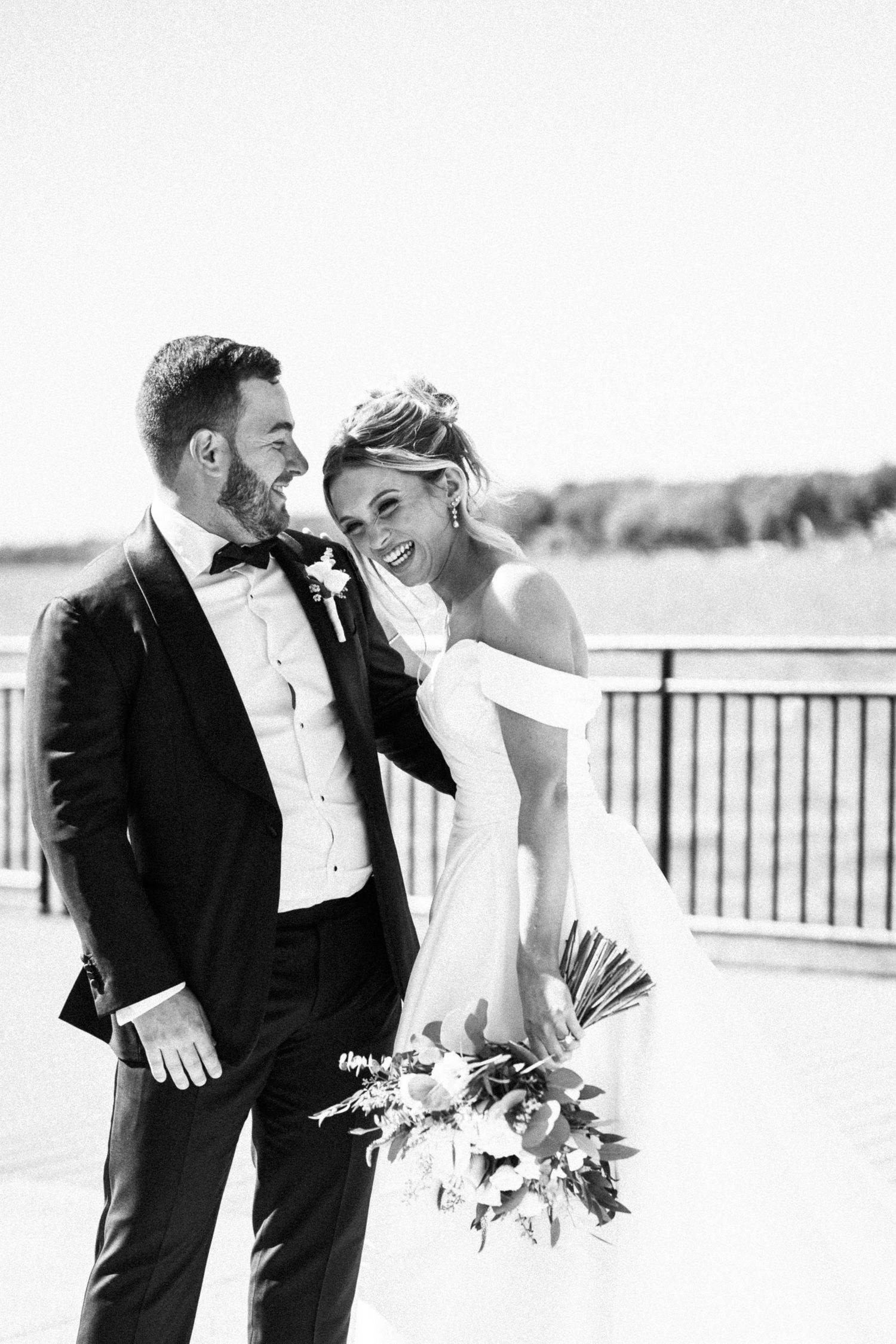 Black and white photo of laughing bride in off-shoulder gown with groom in tuxedo by waterfront.