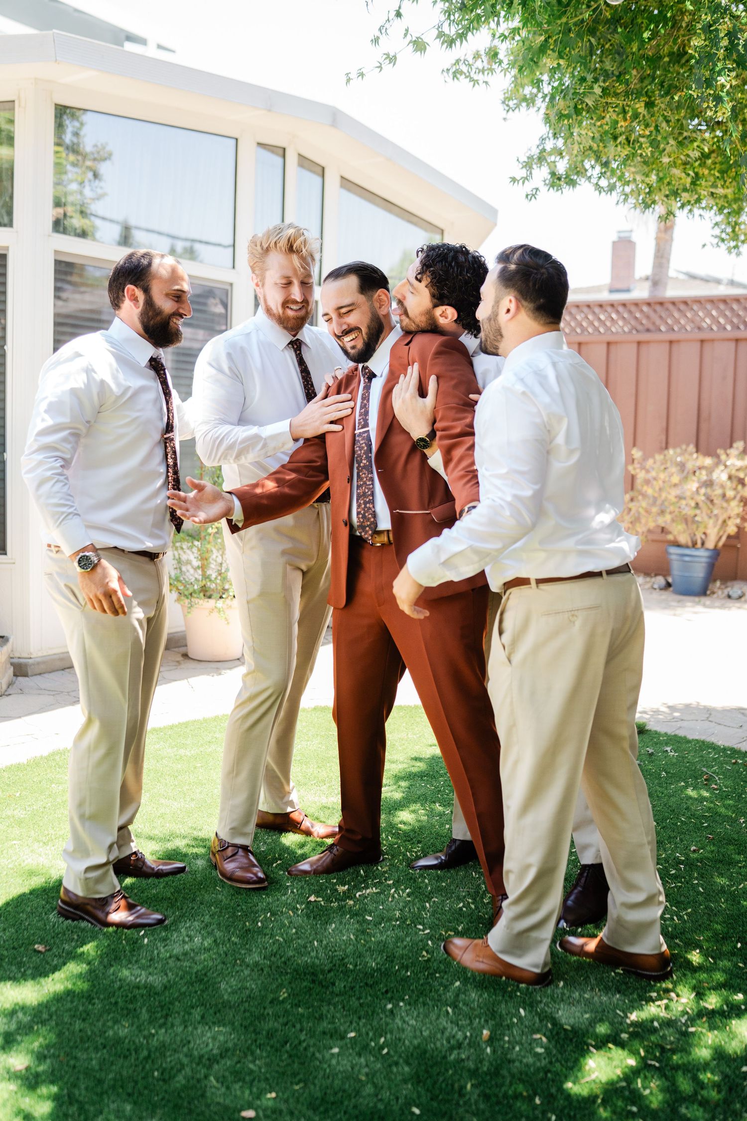 Groom in brown suit surrounded by laughing groomsmen in white shirts on green lawn.