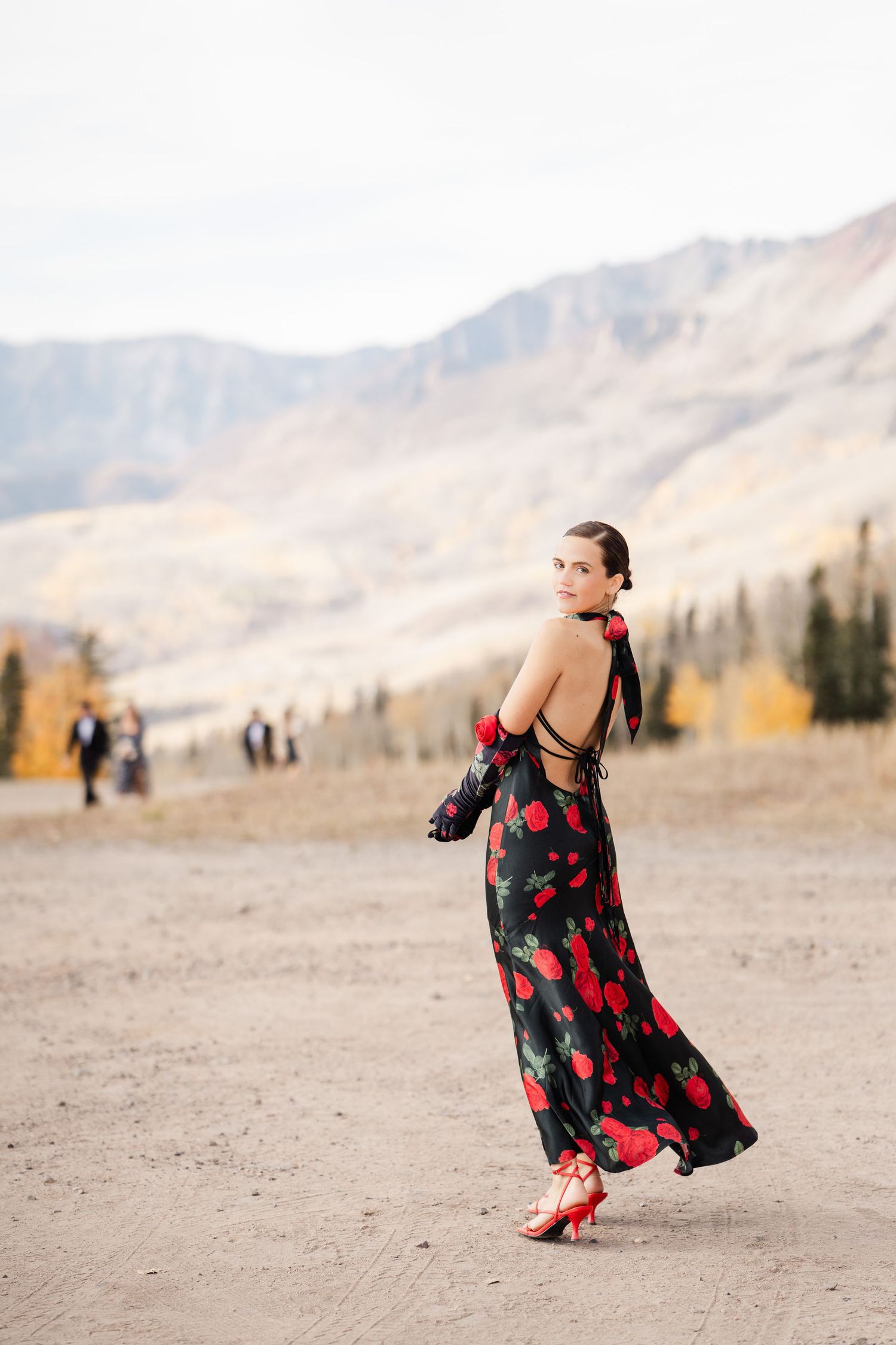Woman in elegant black and red floral backless gown with red heels posing in mountain landscape setting.