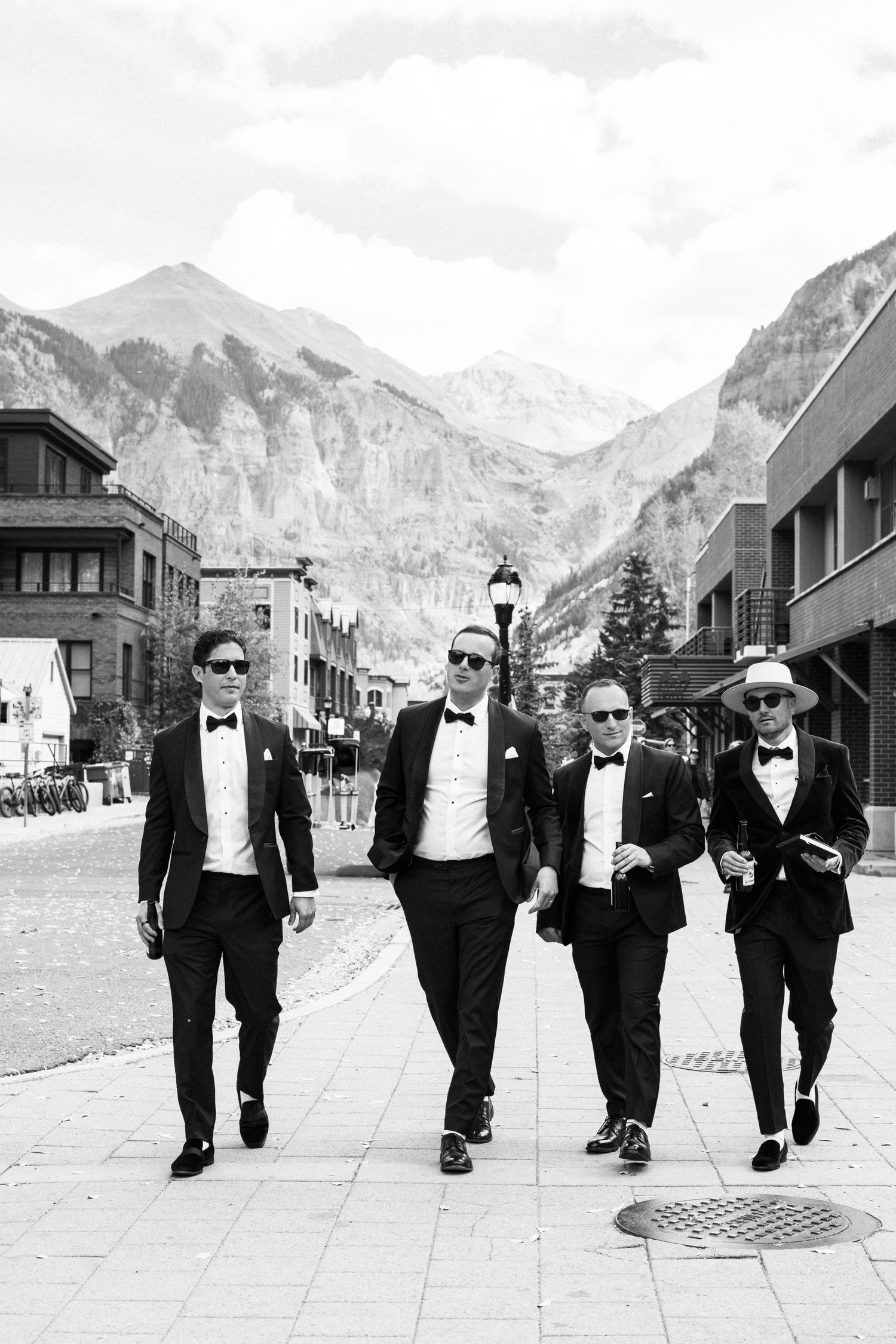 Four groomsmen in black tuxedos walking confidently through mountain town street in black and white photo.