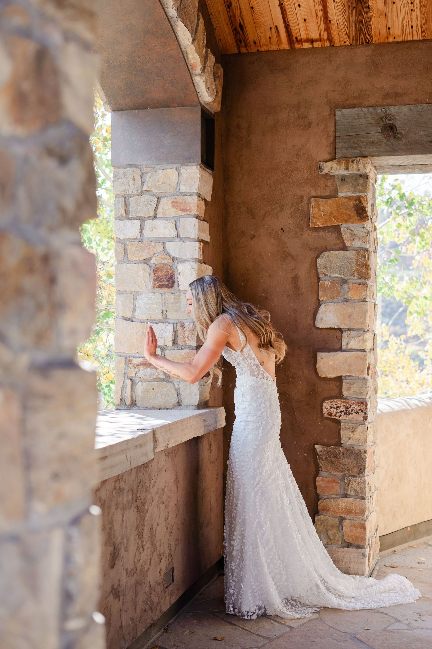 A bride in a lace wedding gown poses against a rustic stone wall in warm natural light.