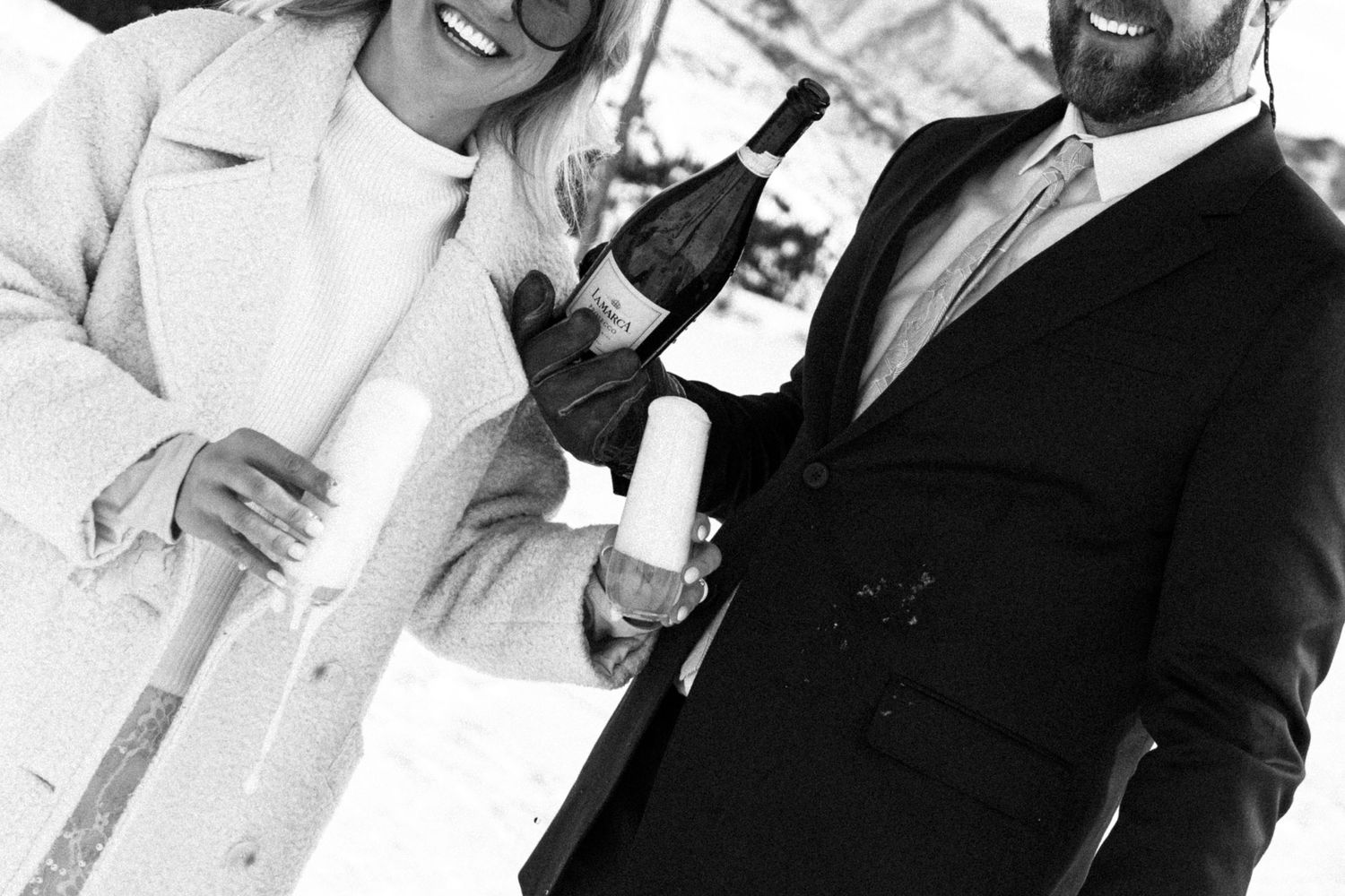 Black and white photo of a smiling couple celebrating with a champagne bottle in snowy setting.