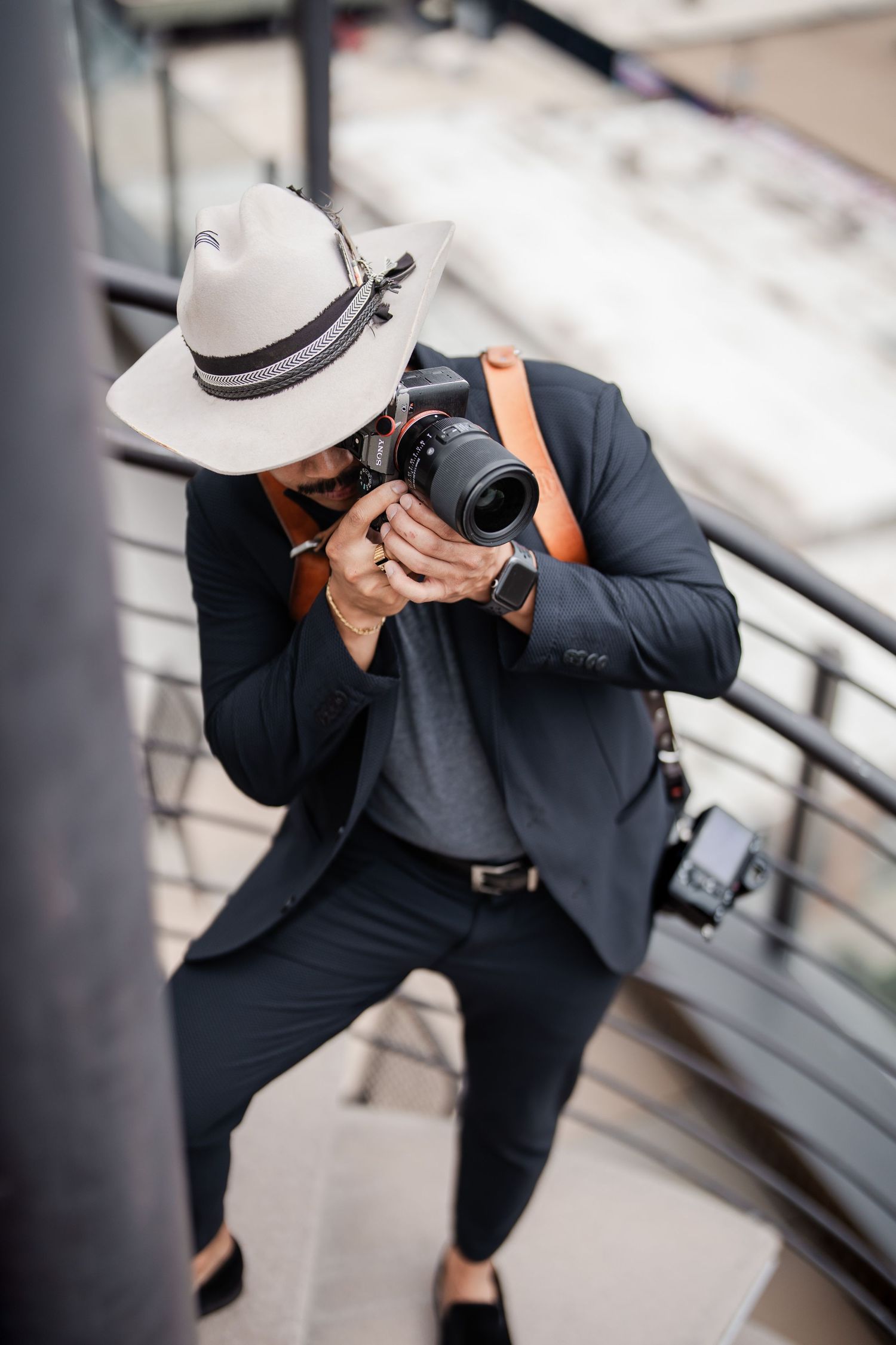 Photographer in white hat and dark suit capturing photos with DSLR camera on outdoor stairs.