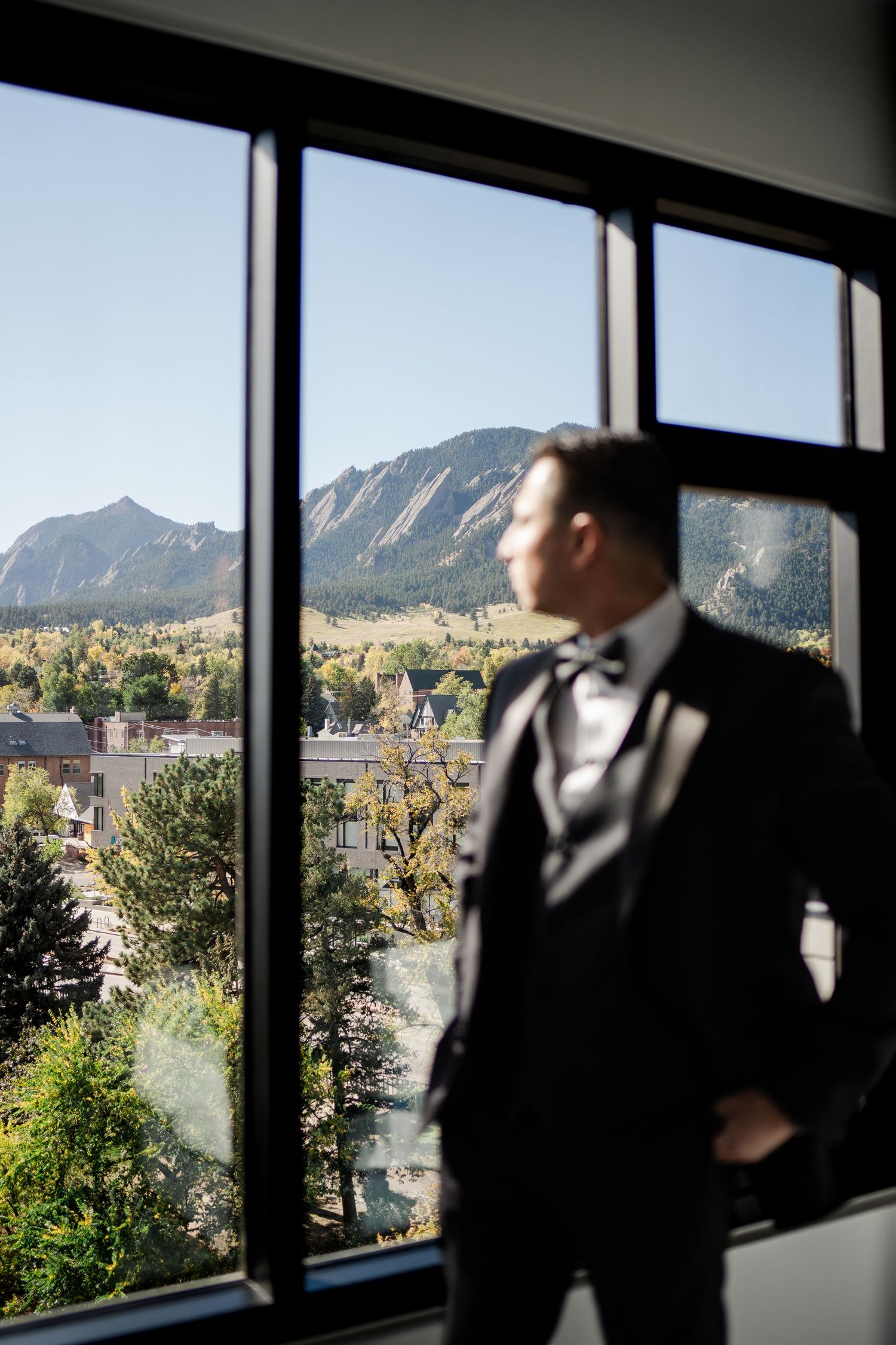 Groom in tuxedo gazing out large window at Boulder Flatirons mountains on wedding day.