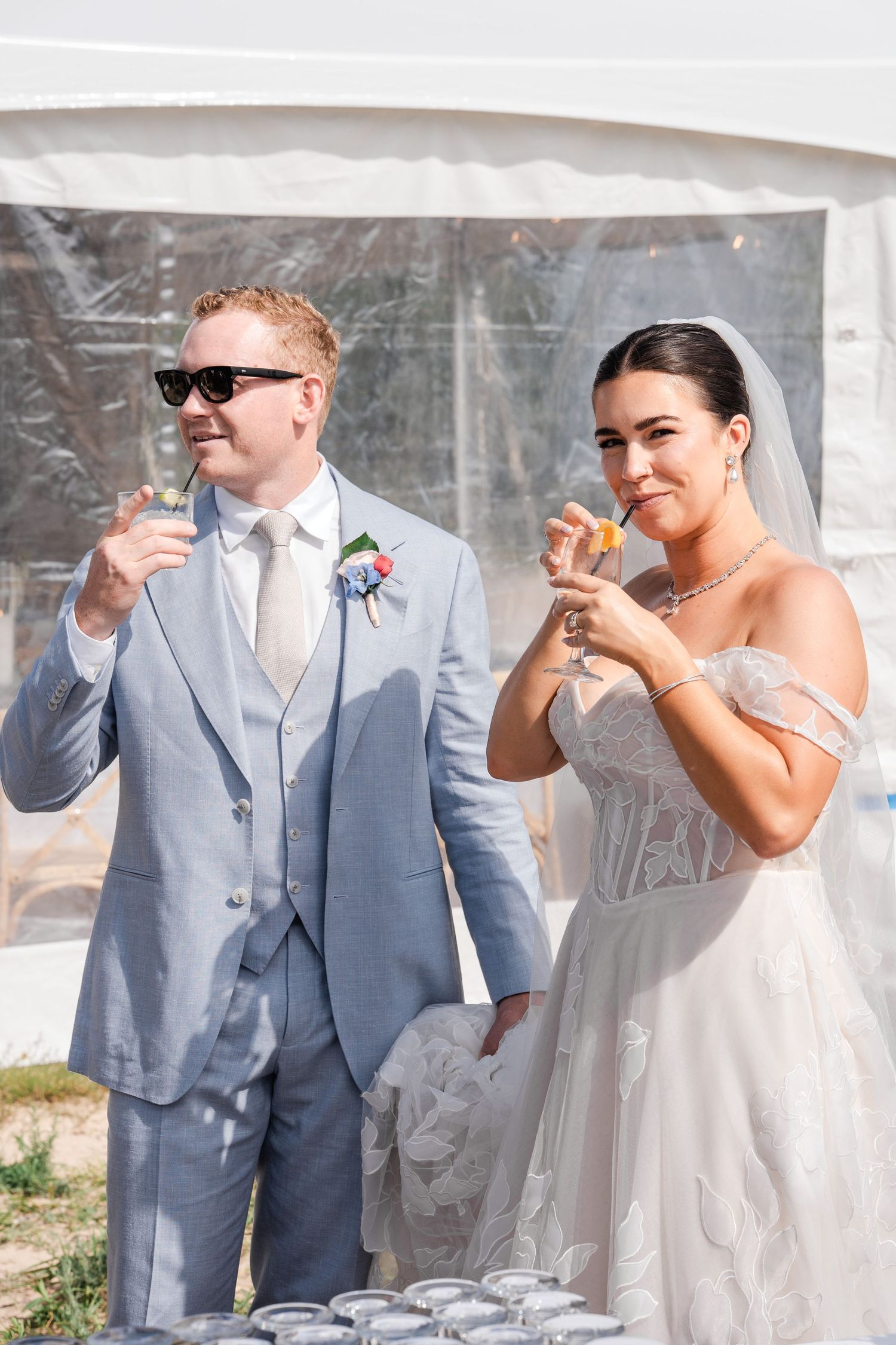 Bride and groom toast champagne under white tent at outdoor summer wedding reception on sunny day.