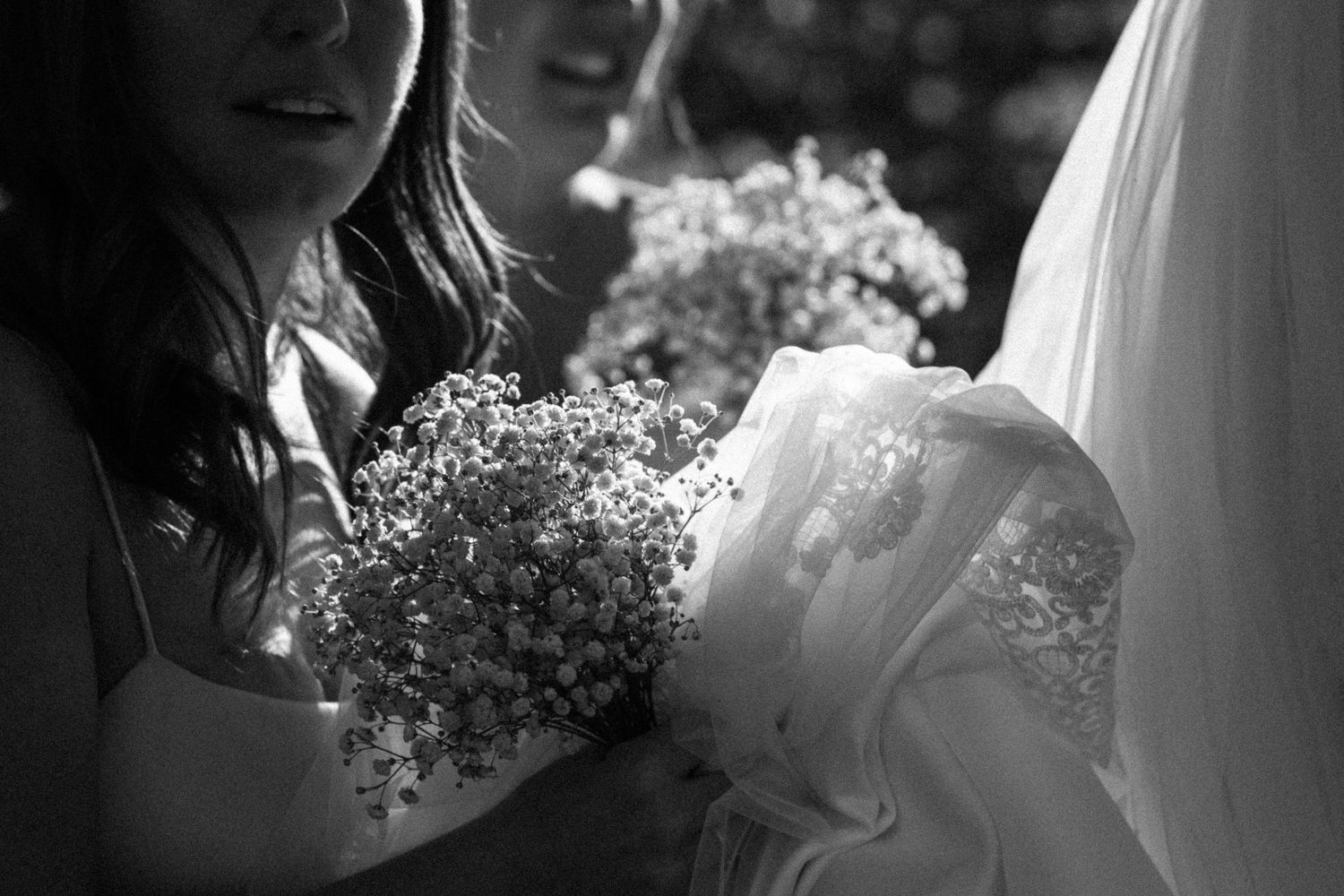 Black and white close-up of bridesmaid holding baby's breath bouquet beside bride's flowing white wedding gown.