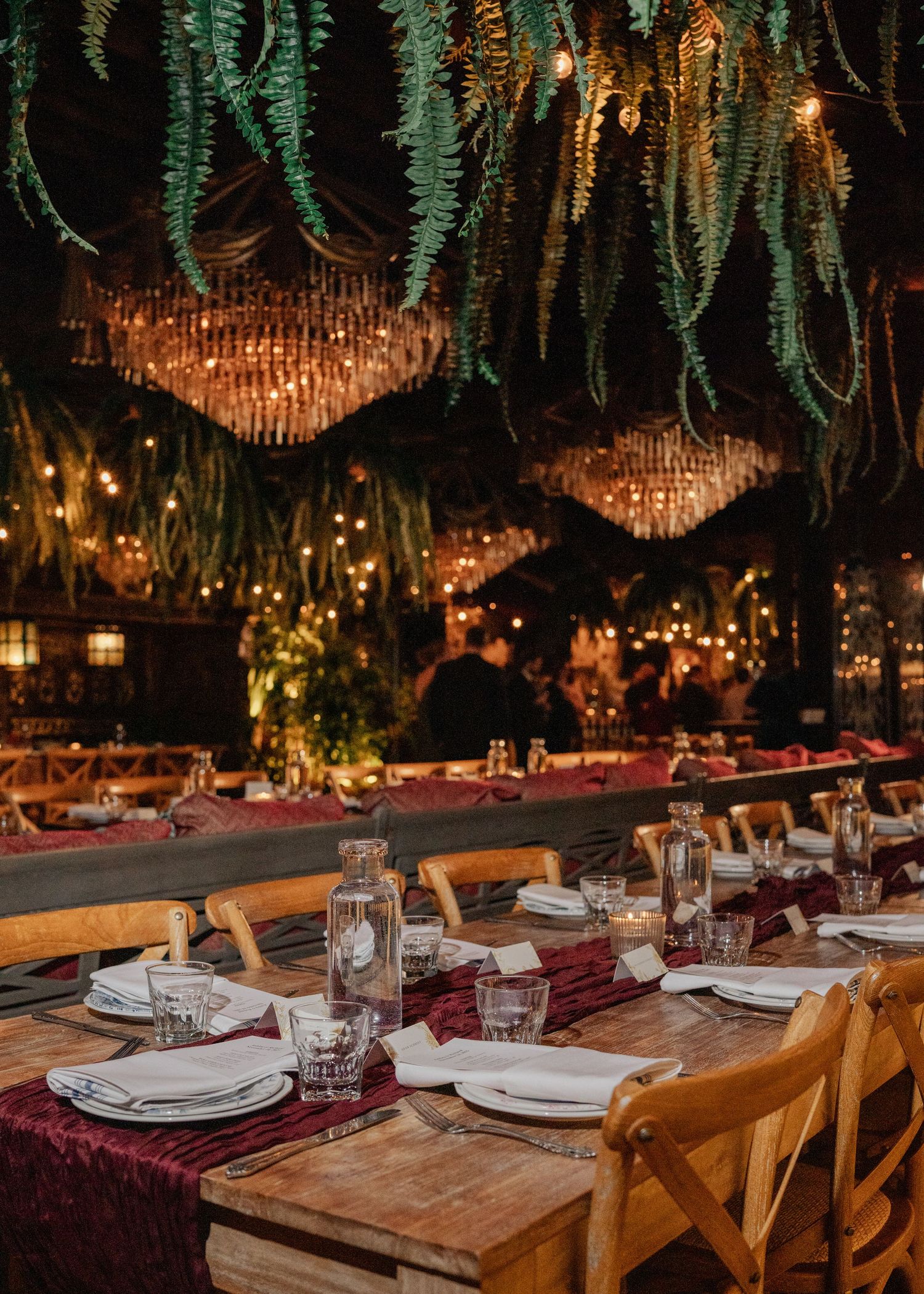 Elegant wedding reception with long wooden tables, burgundy velvet runners, hanging ferns, and warm chandelier lighting.