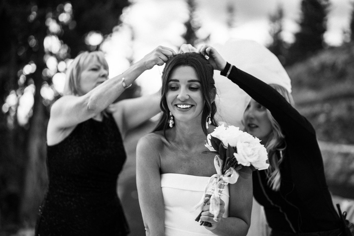 Bridesmaids help smiling bride adjust her veil outdoors in black and white wedding photo.