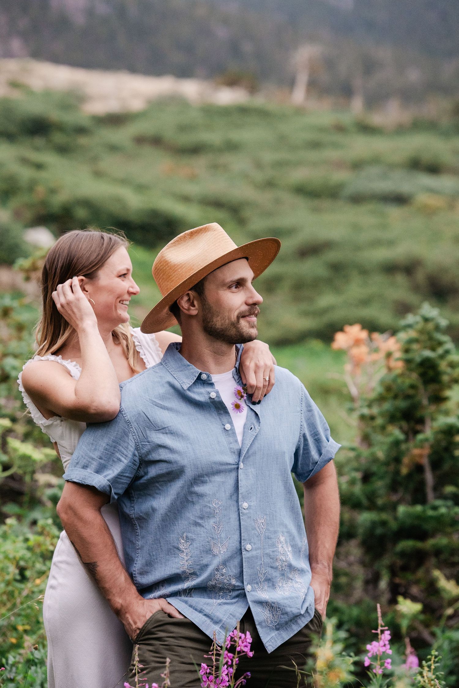 Engaged couple posing together in alpine meadow with wildflowers, woman in white dress leaning on mans shoulder.