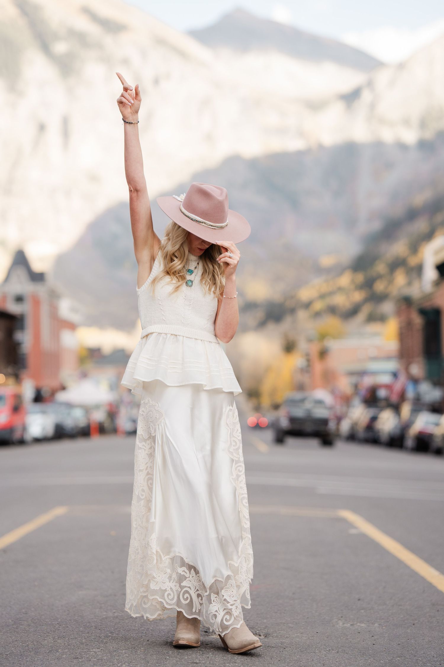 Boho bride in white lace dress and pink felt hat raises hand joyfully on a mountain town street in autumn.