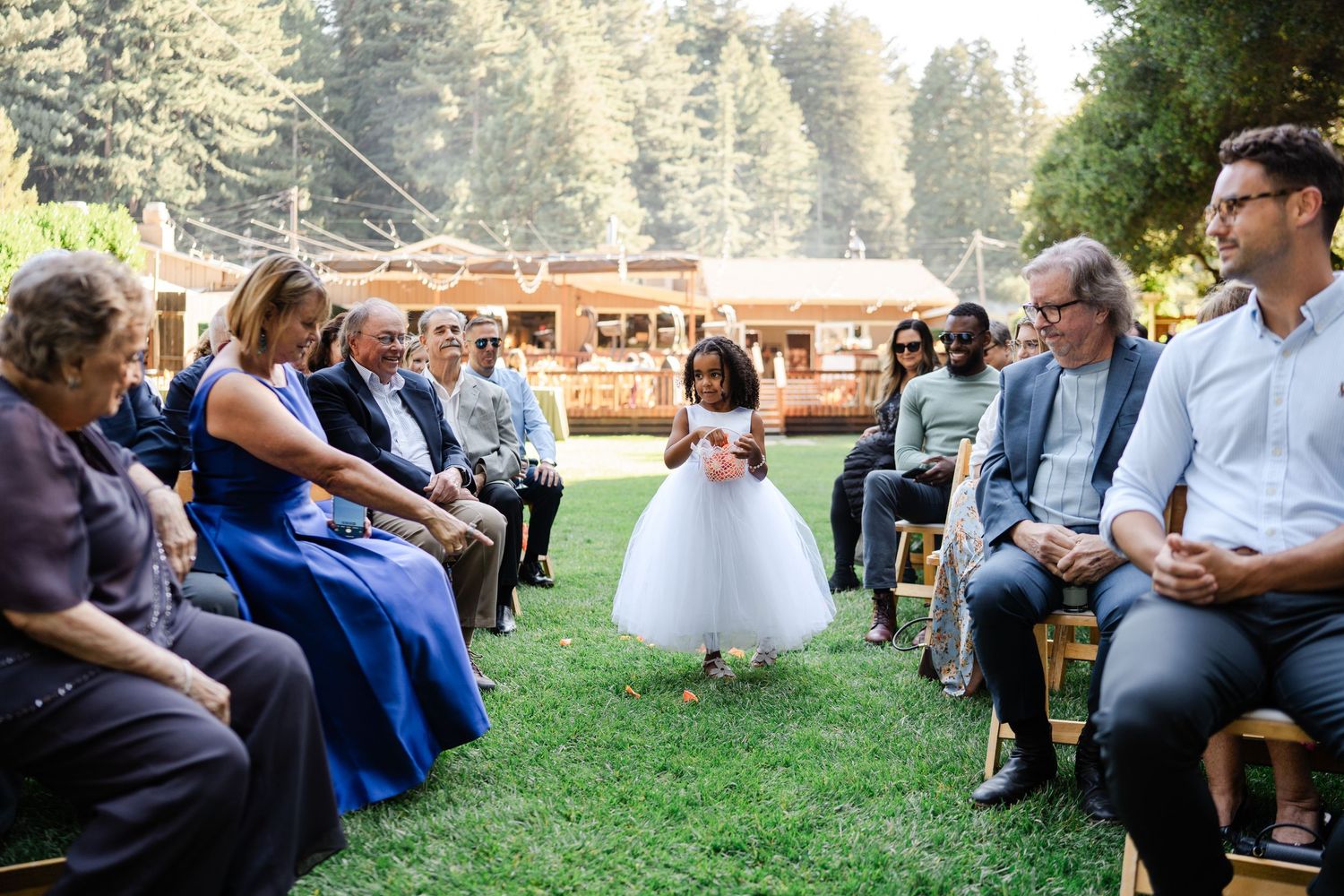 Adorable flower girl in white tutu dress walking down outdoor wedding ceremony aisle.