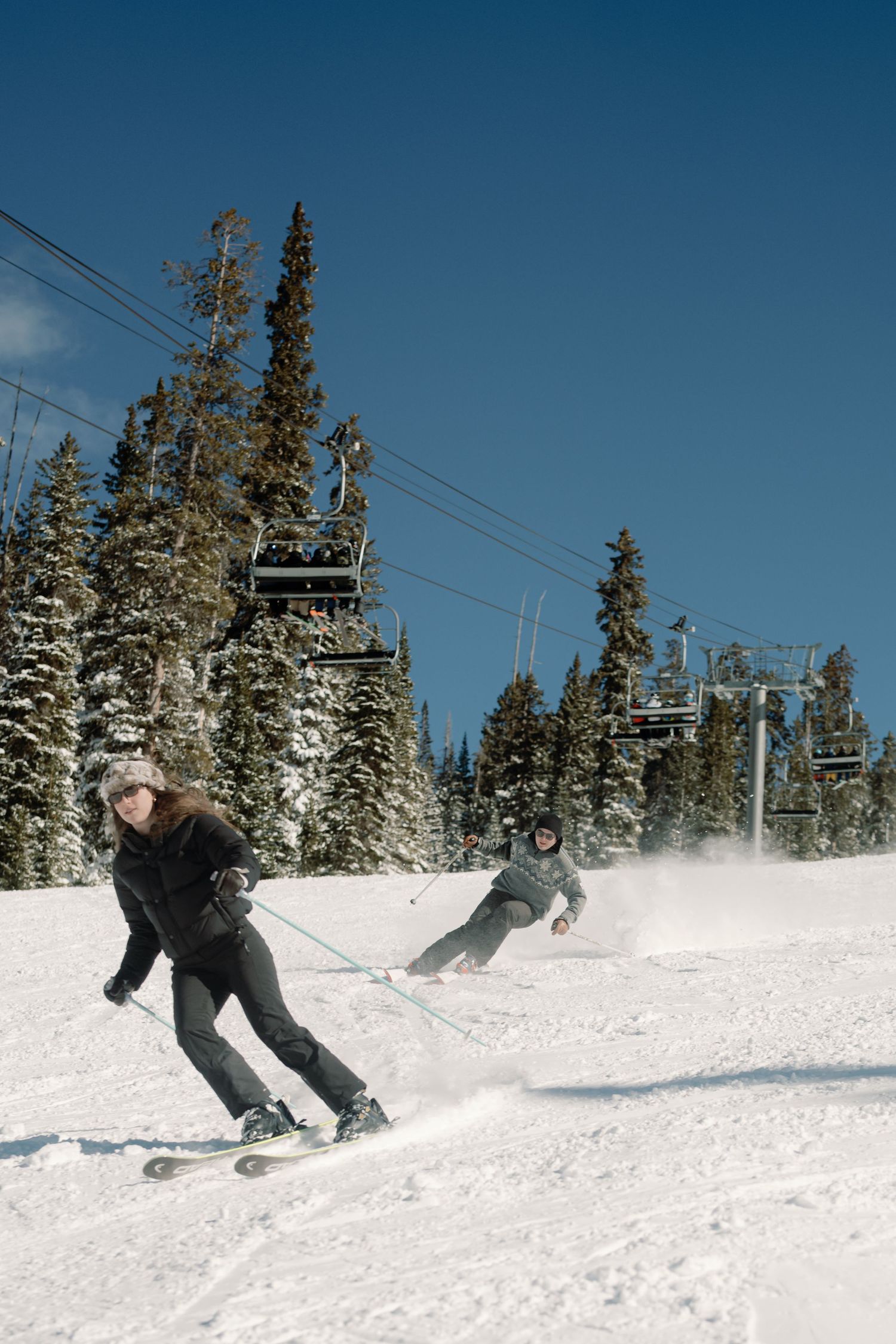 Two skiers racing downhill on groomed slope with ski lift and blue sky above.
