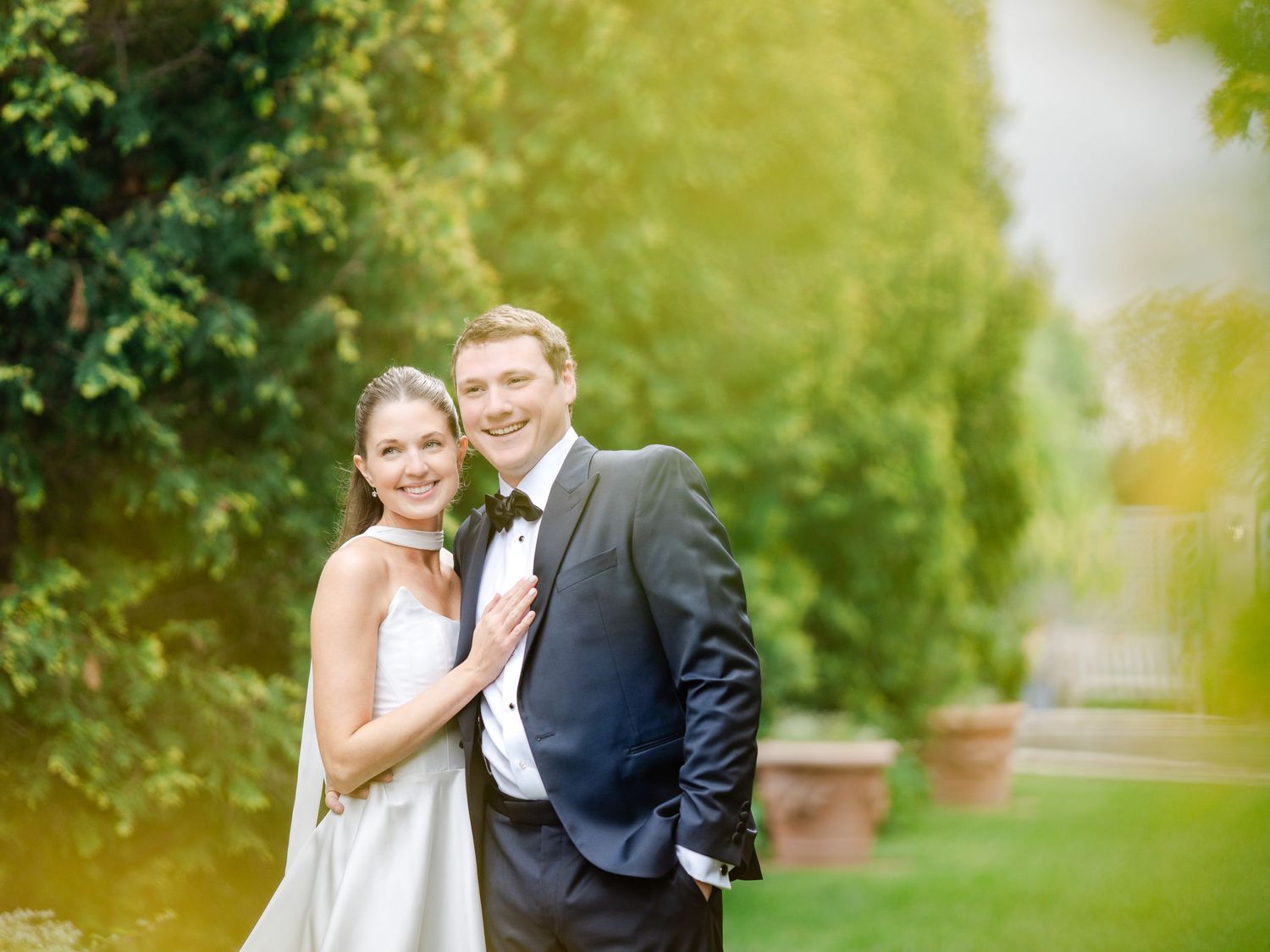 A smiling bride in a white gown and groom in a dark tuxedo pose together in a lush garden setting.