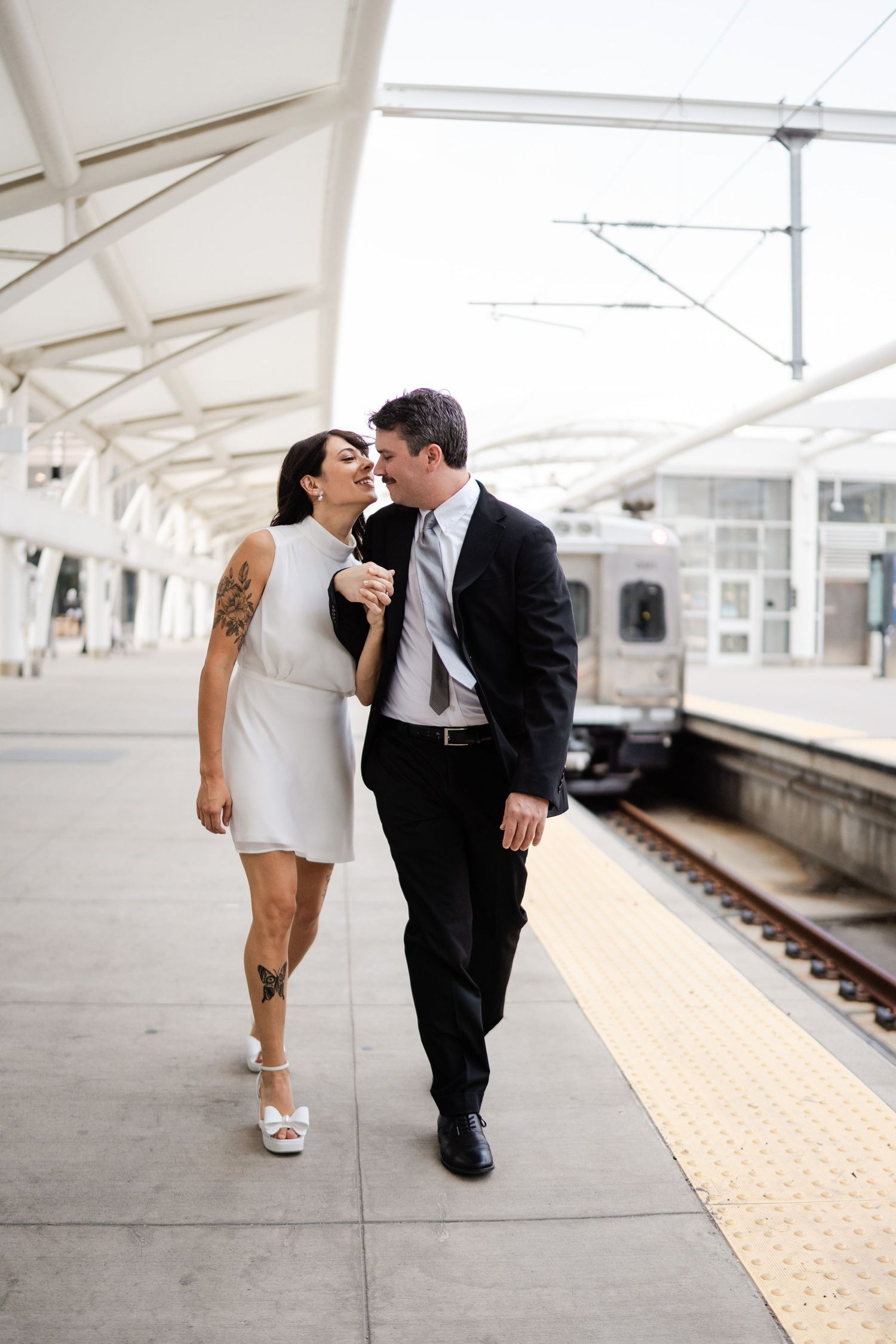 Bride and groom in white and black attire walking and laughing together on a train station platform.