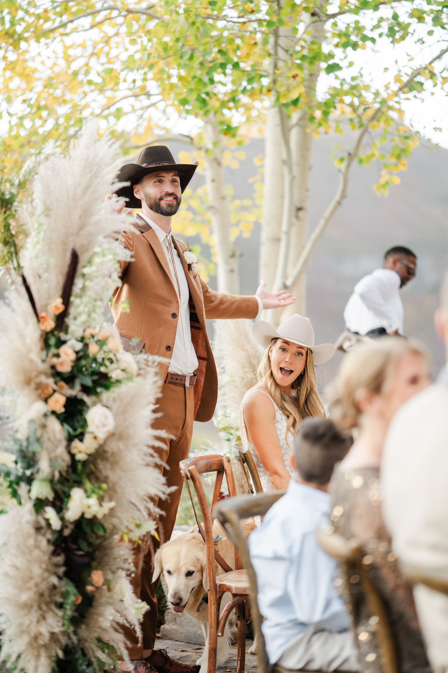 A joyful couple in cowboy hats celebrates their western-themed wedding reception with their dog nearby.