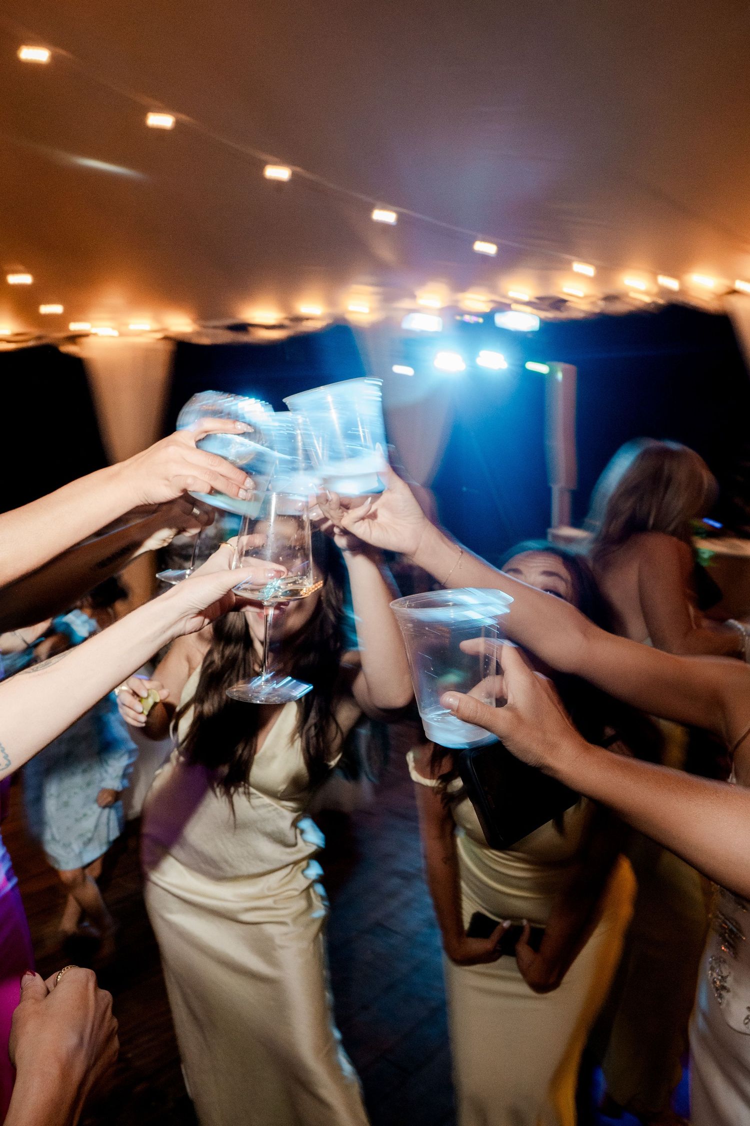 Wedding guests raising glasses in a lively toast on the dance floor under string lights.
