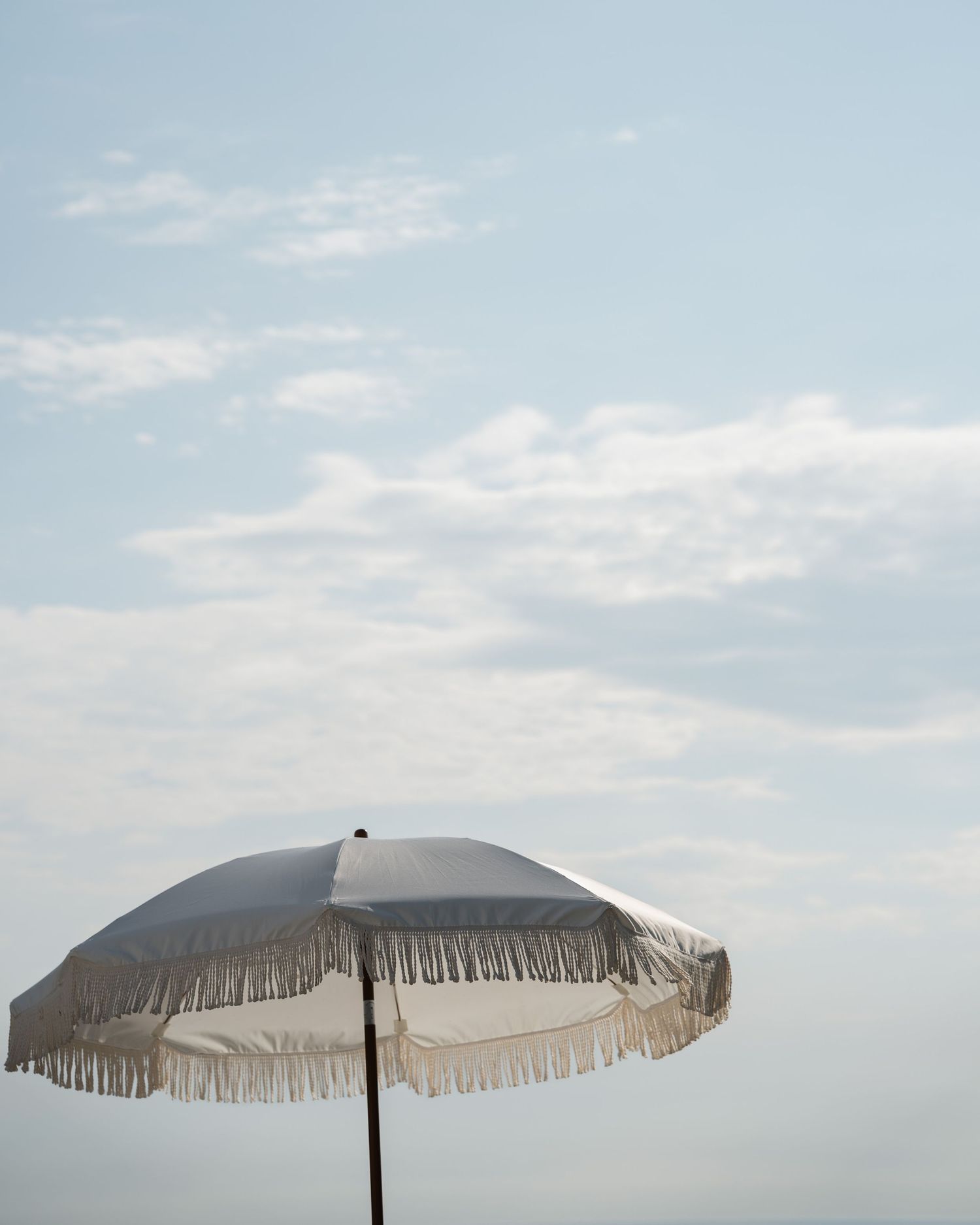White fringed beach umbrella against a soft blue cloudy sky at an outdoor wedding.