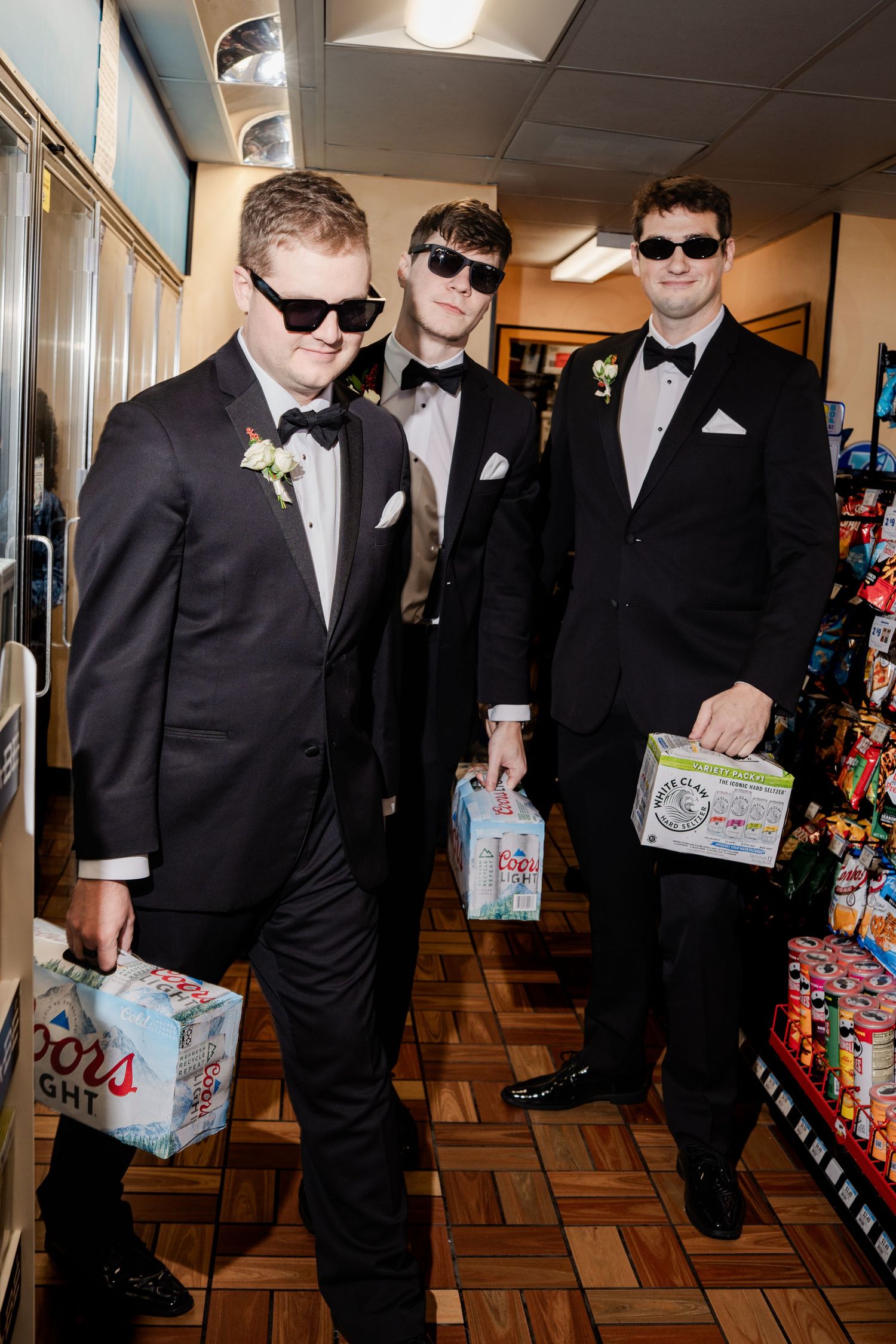 Three groomsmen in tuxedos and sunglasses holding beer cases inside a convenience store.