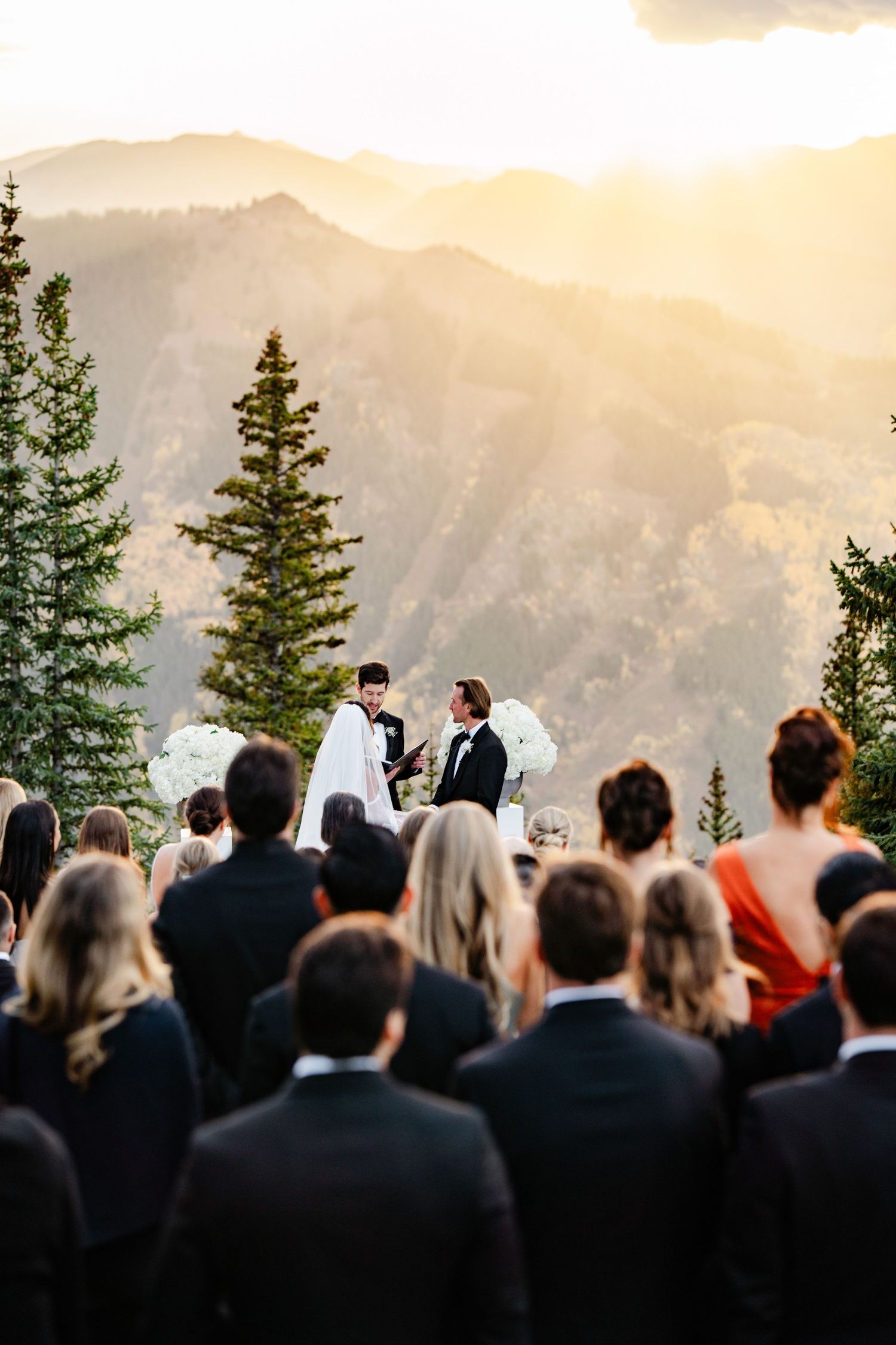 Couple exchanging vows at a golden sunset mountain wedding ceremony with guests seated outdoors.