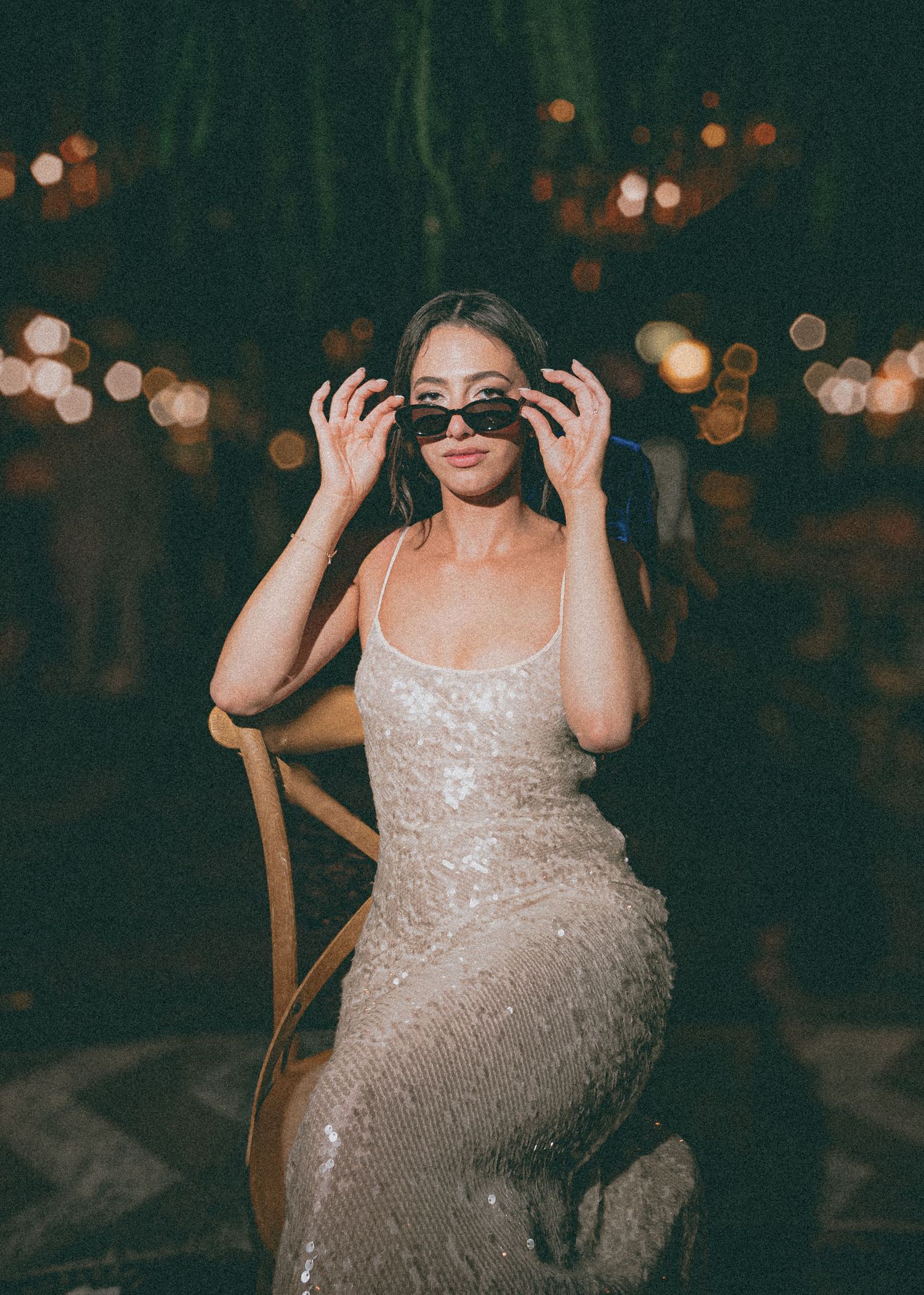 Bride in sequined gown wearing sunglasses seated on a wooden chair with bokeh lights in the background.