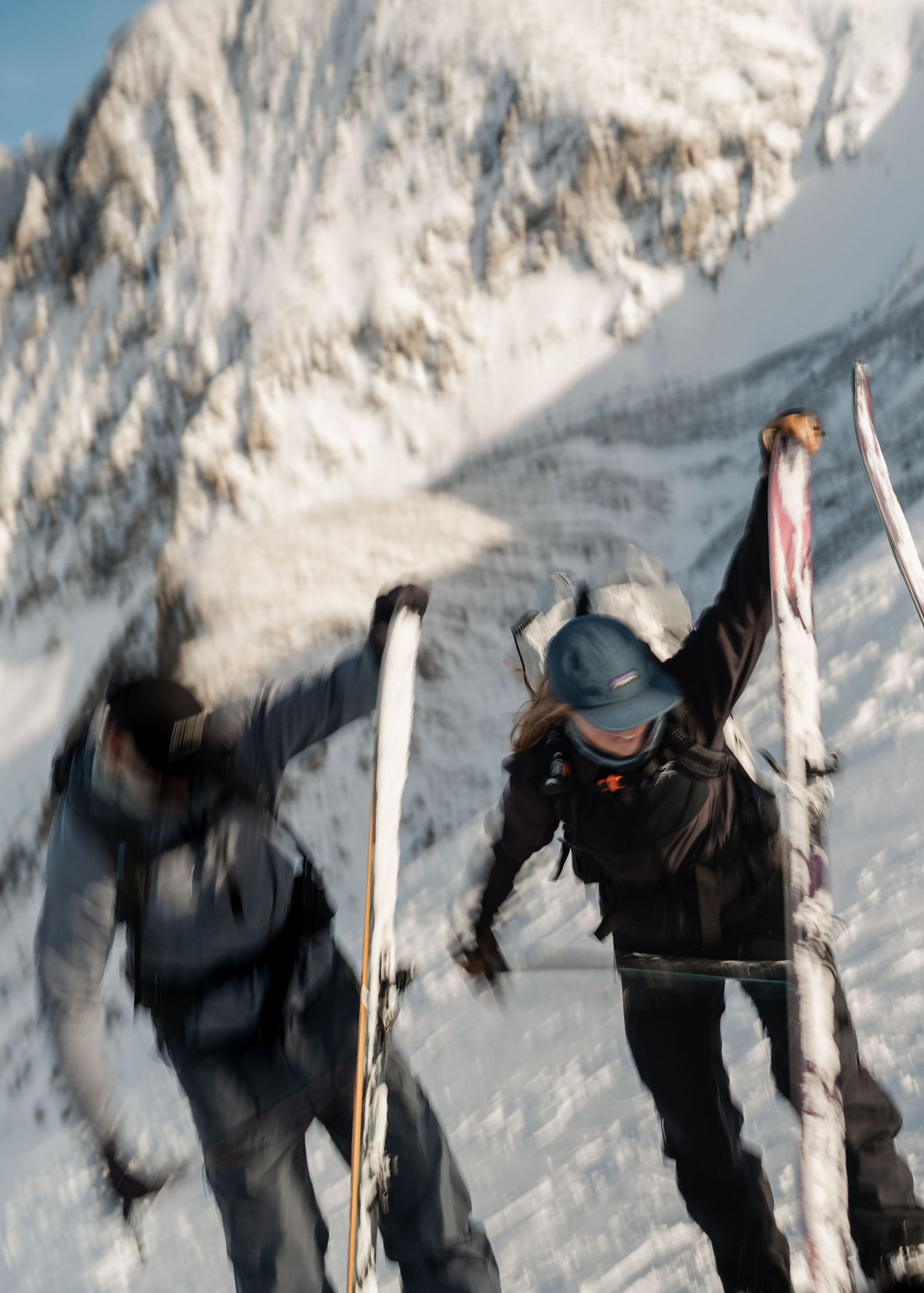 Two skiers carry their skis while hiking up a steep snow-covered mountain face in a motion-blurred action shot.