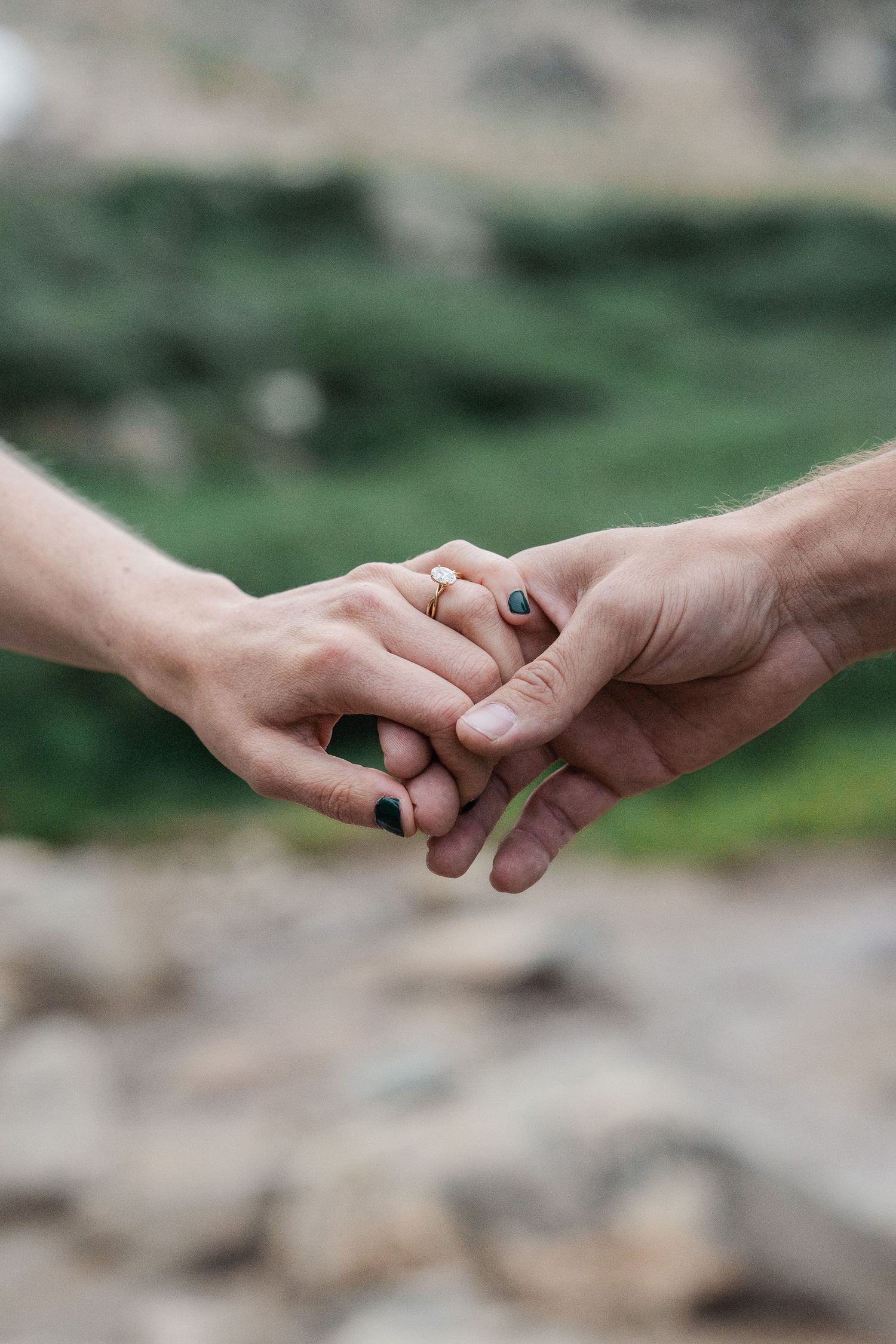 Close-up of two hands holding gently, one wearing a delicate engagement ring, with a blurred green mountain backdrop.