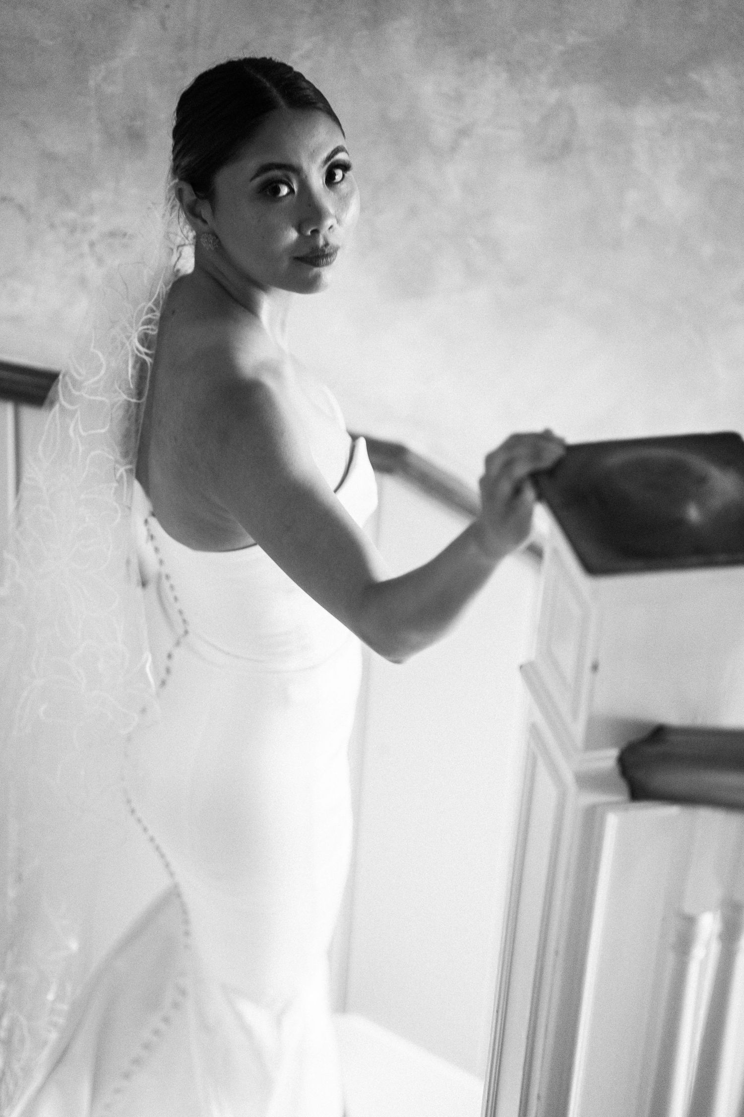 Elegant bride in fitted white gown and veil poses in black and white bridal portrait indoors.