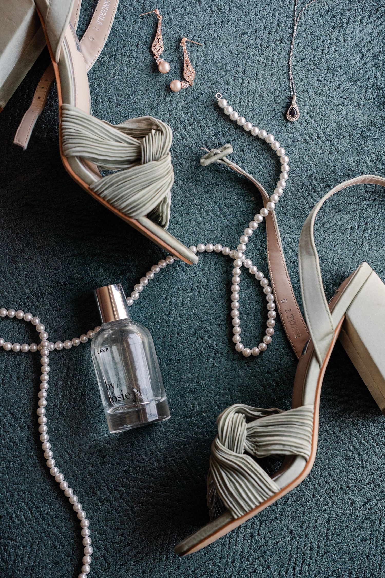 Sage green knotted heels with pearl necklace, perfume bottle, and delicate earrings on teal carpet.