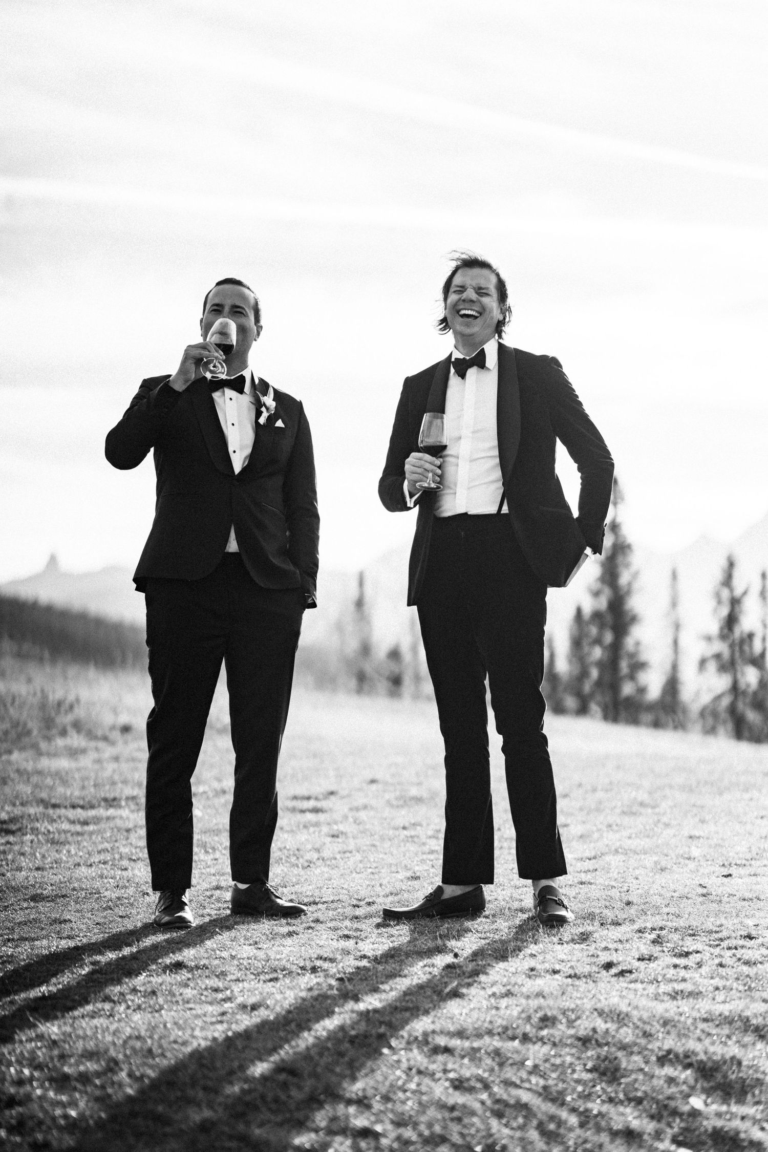 Two groomsmen in black tuxedos laughing and drinking outdoors in black and white wedding photo.