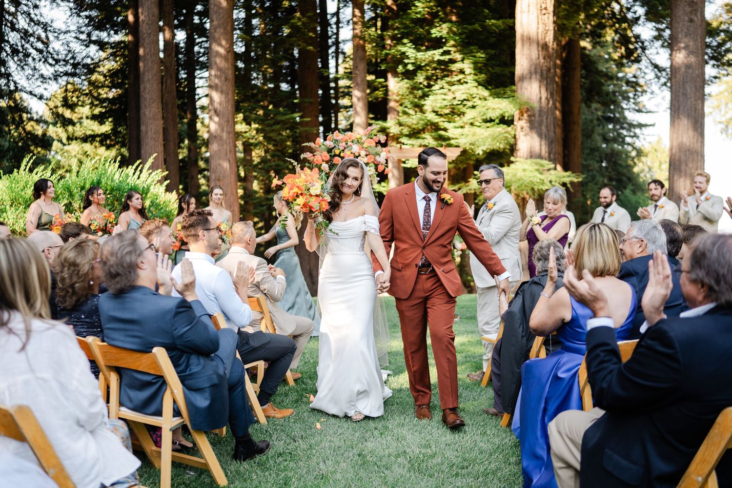 Bride and groom walking down the aisle through redwood forest as guests applaud their outdoor ceremony.