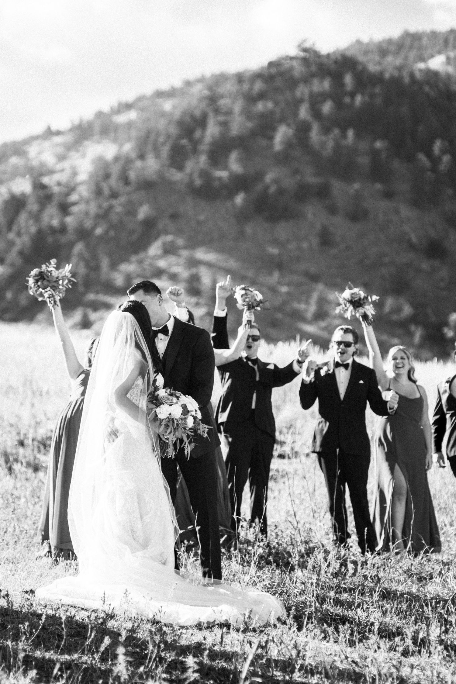 Black and white photo of bride and groom kissing while wedding party celebrates in mountain meadow.