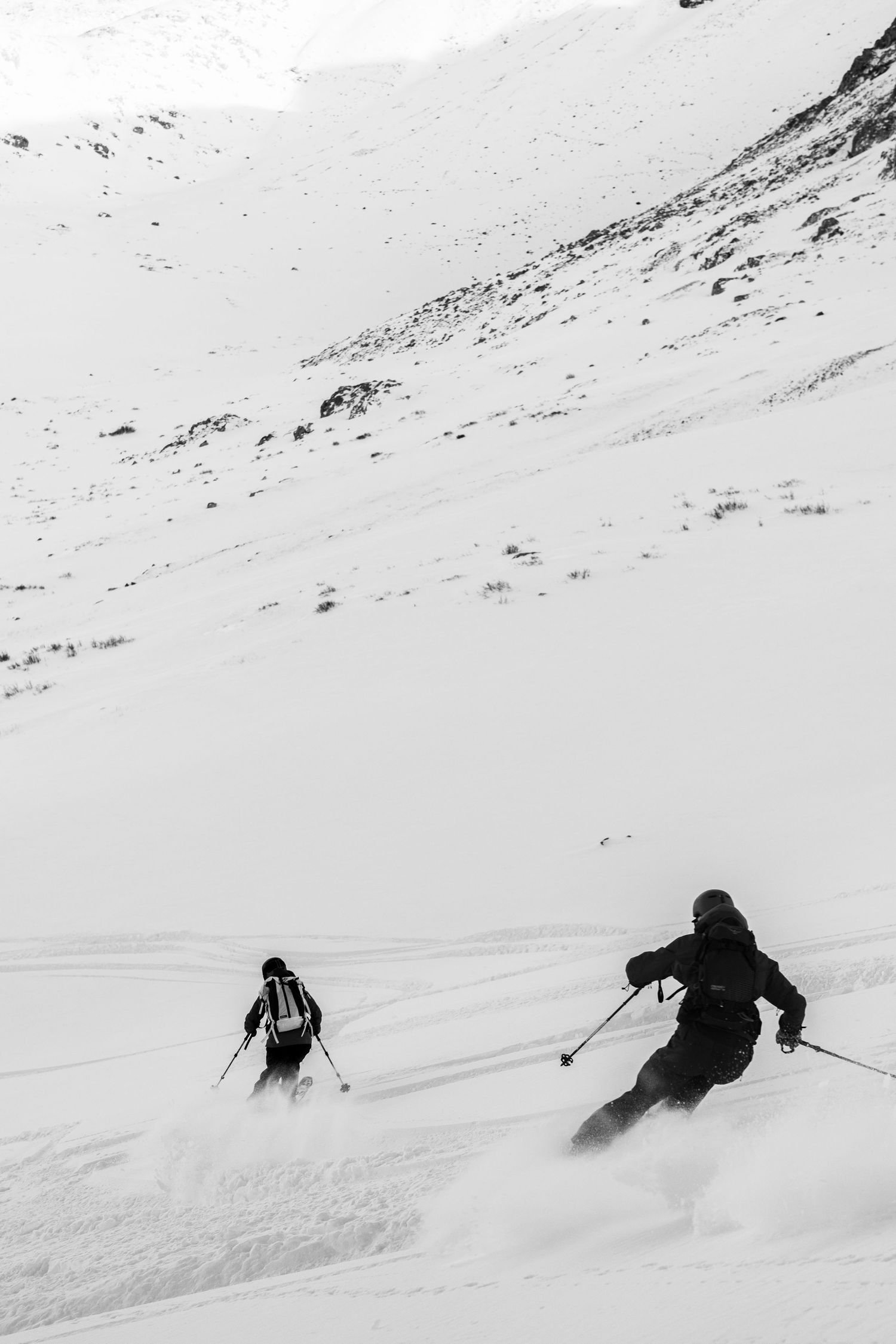 Black and white action shot of two skiers descending steep powder slopes in mountain backcountry.
