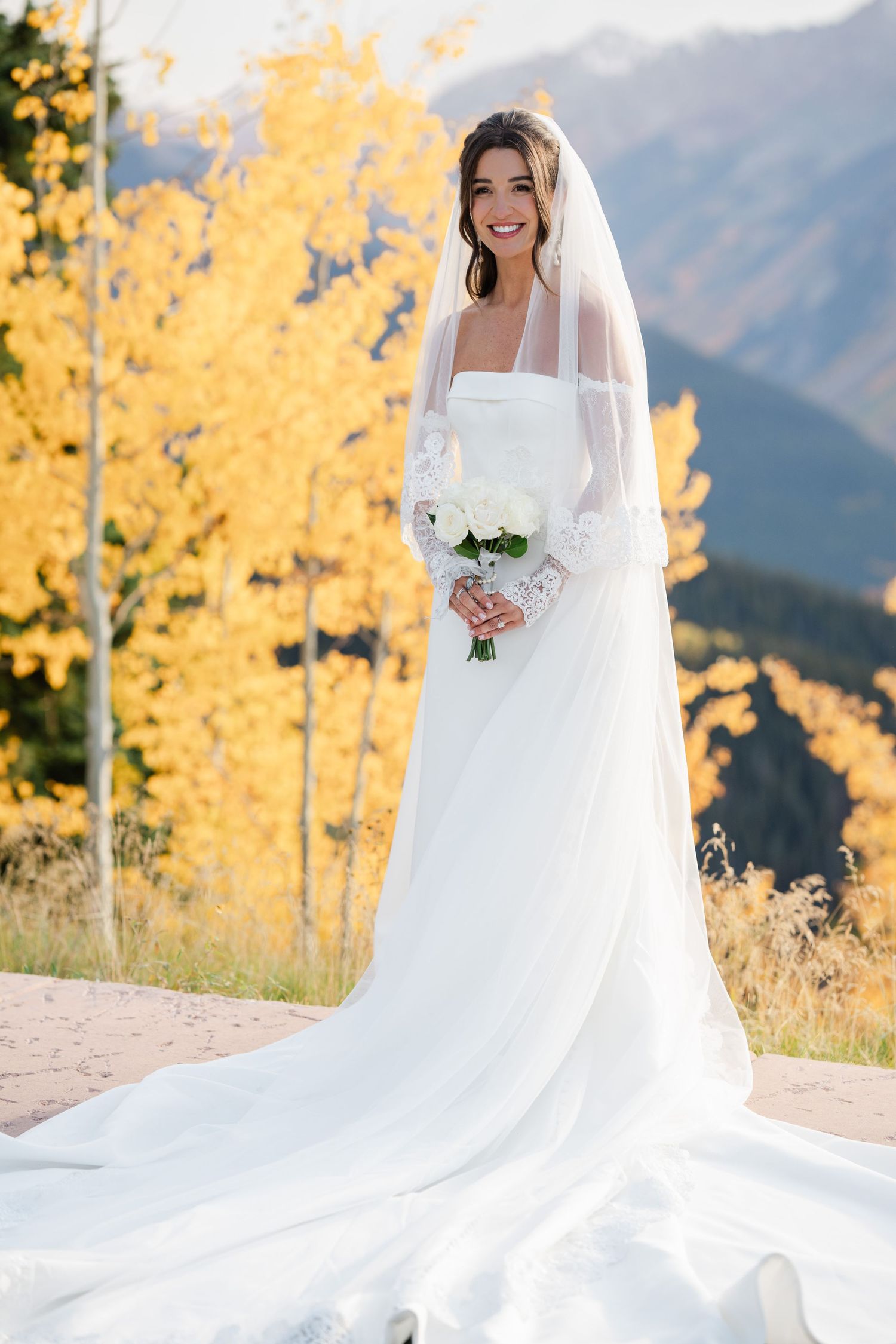 Radiant bride in white gown with long veil holding bouquet surrounded by golden fall foliage and mountains.