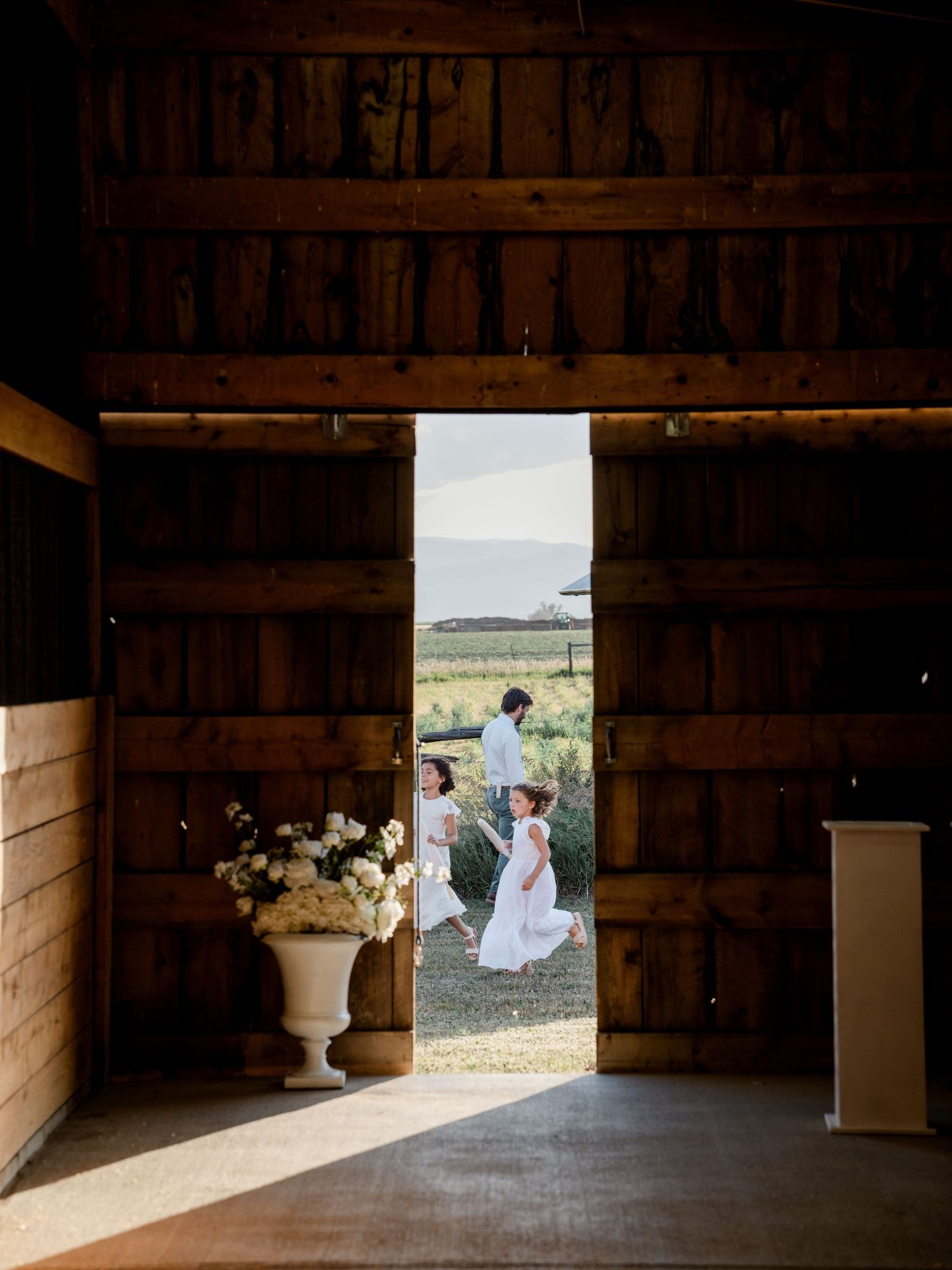 View through rustic barn doors of flower girls playing outside in golden sunlight.