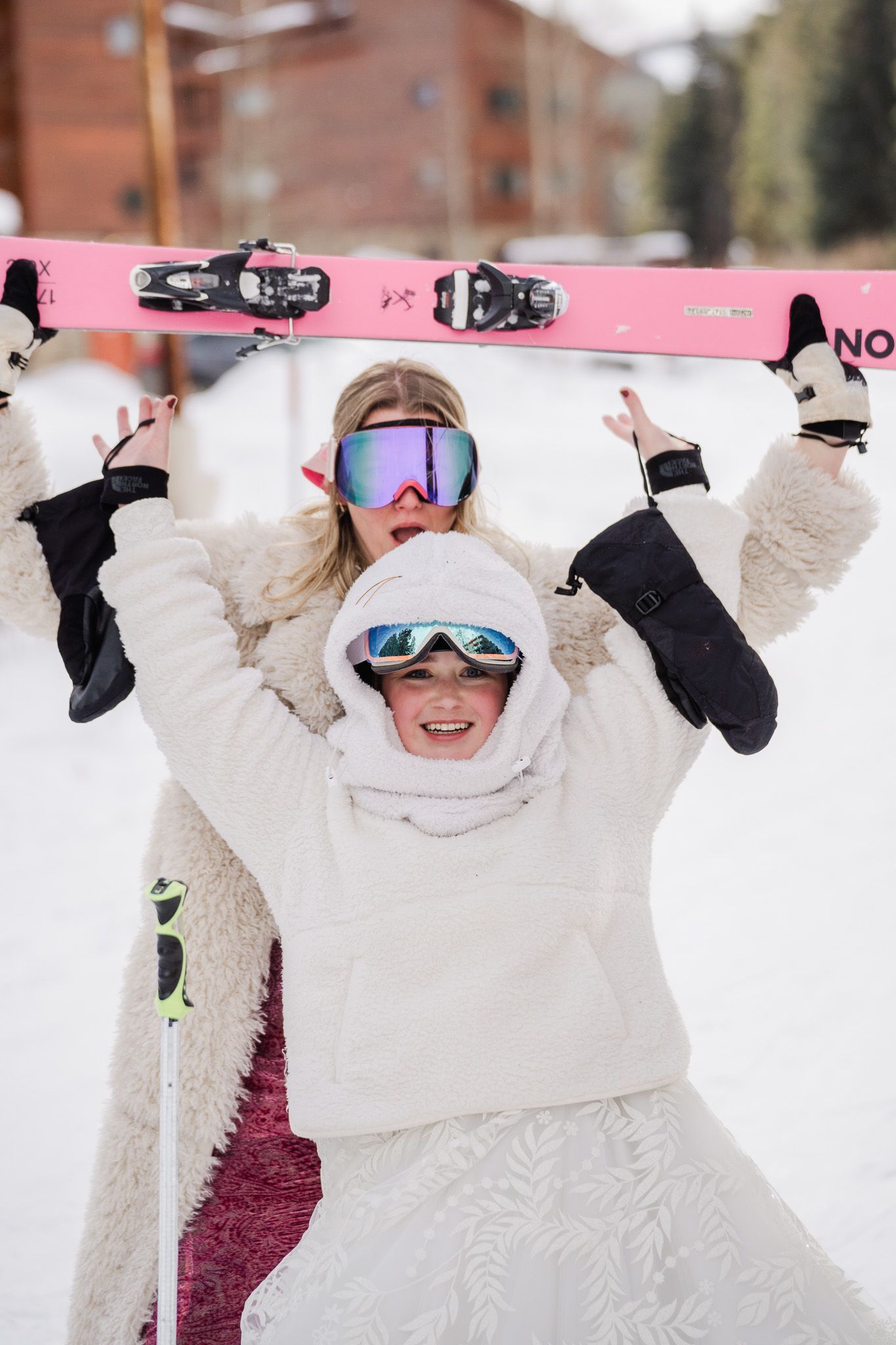 Two women in white winter outfits and ski goggles hold pink skis overhead at a snowy mountain resort.