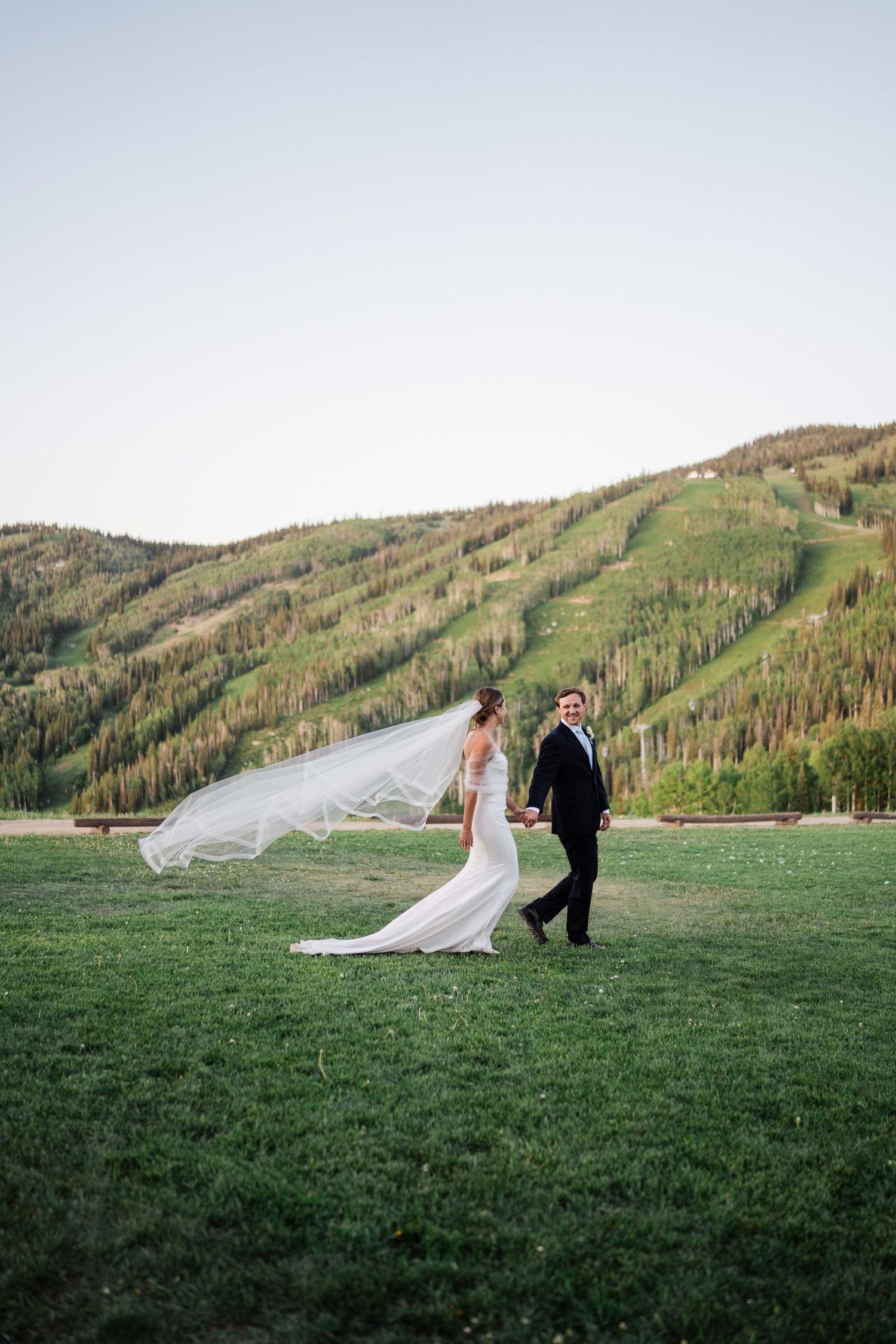 Bride in flowing gown with veil blowing in wind walks with groom on green mountain meadow.