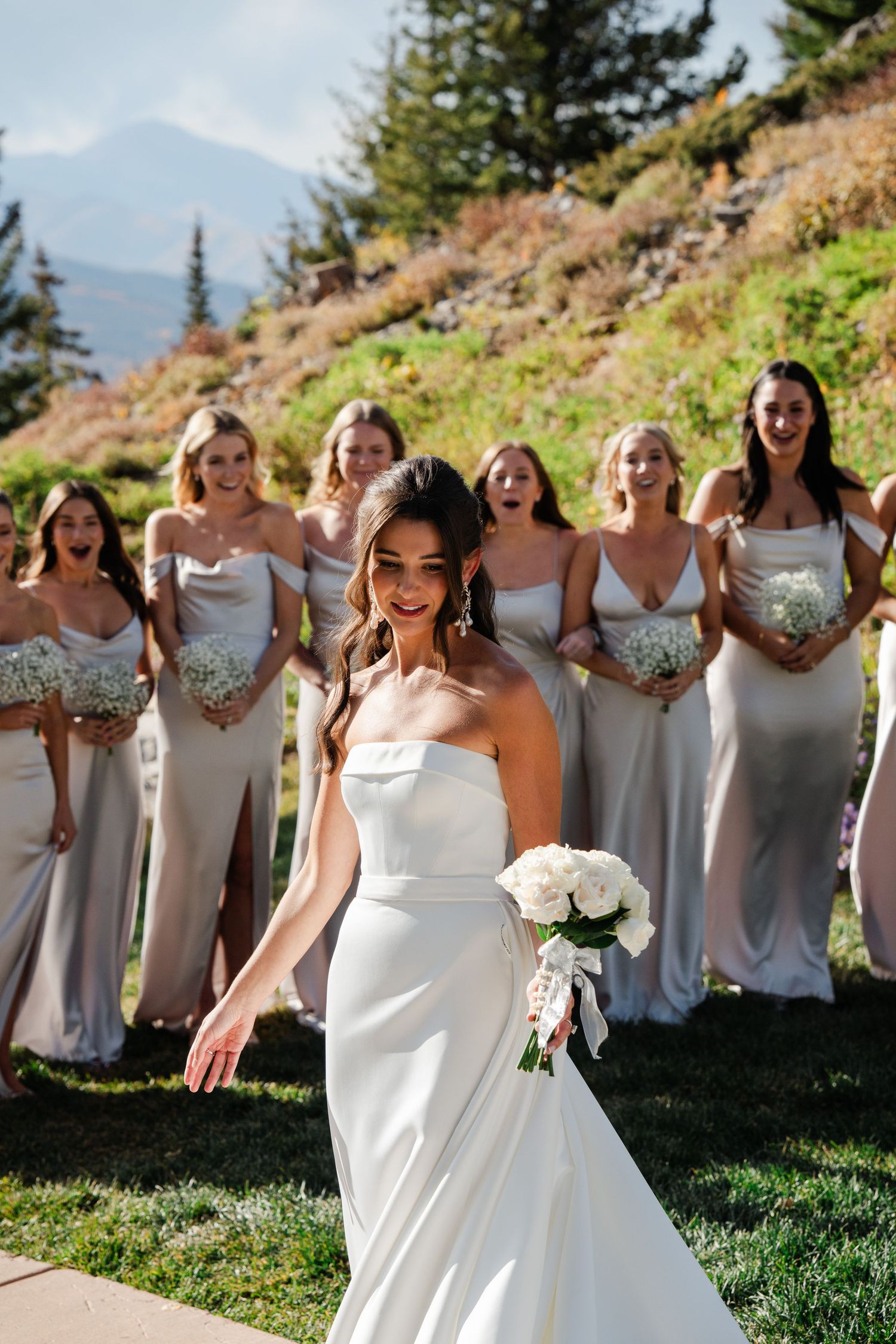 Bride in white strapless gown holds bouquet while excited bridesmaids in champagne dresses react behind her outdoors.