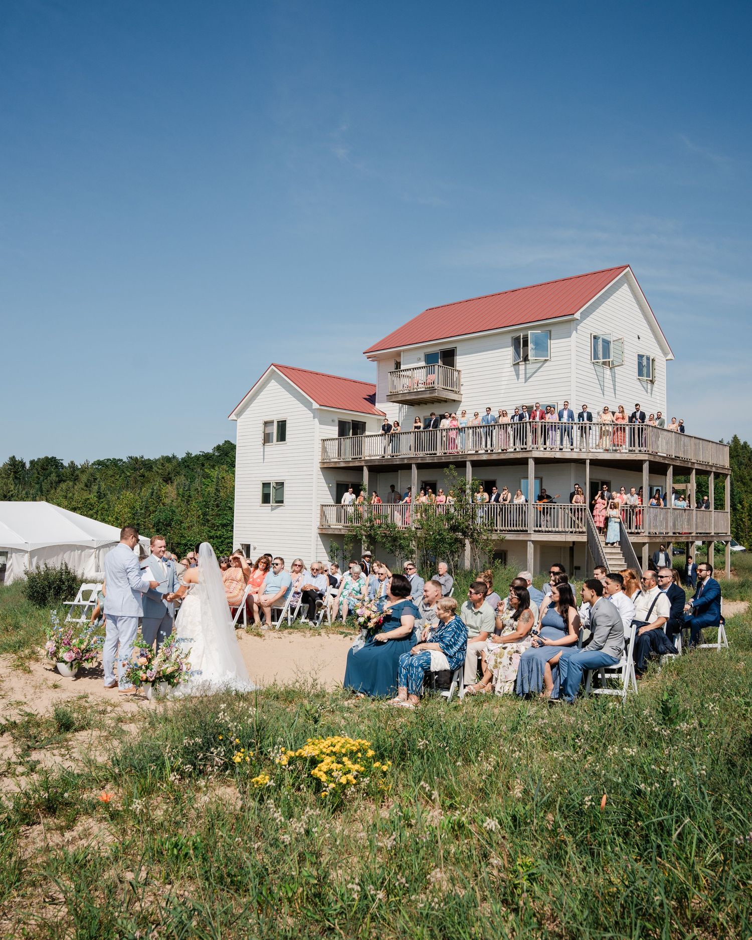Outdoor wedding ceremony on sandy dunes with guests seated and standing on a multi-deck white house with red roof.