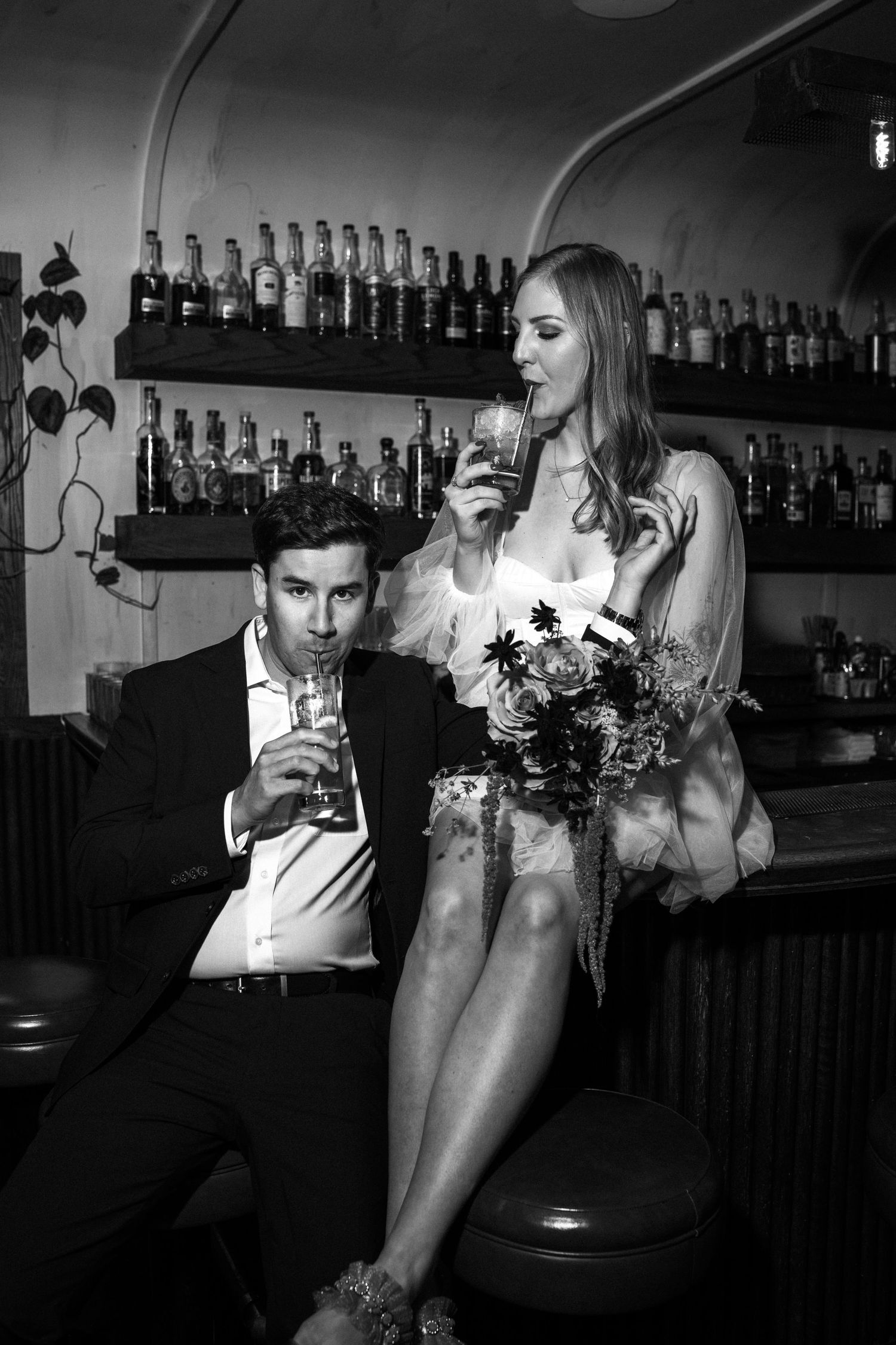 Black and white photo of couple drinking cocktails together at a bar with bottle-lined shelves.