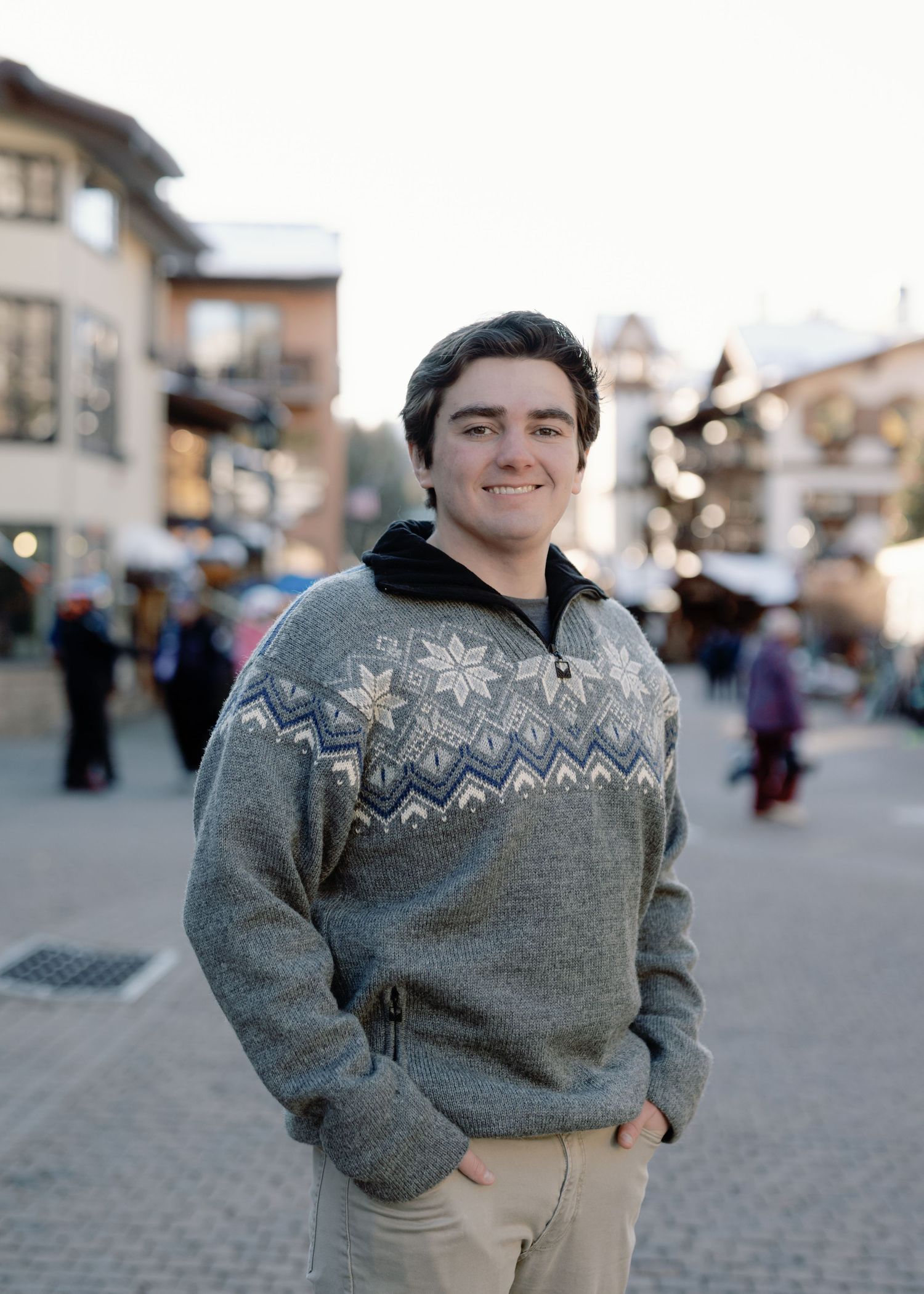 Young man in Nordic patterned sweater smiling in a charming winter mountain village setting.