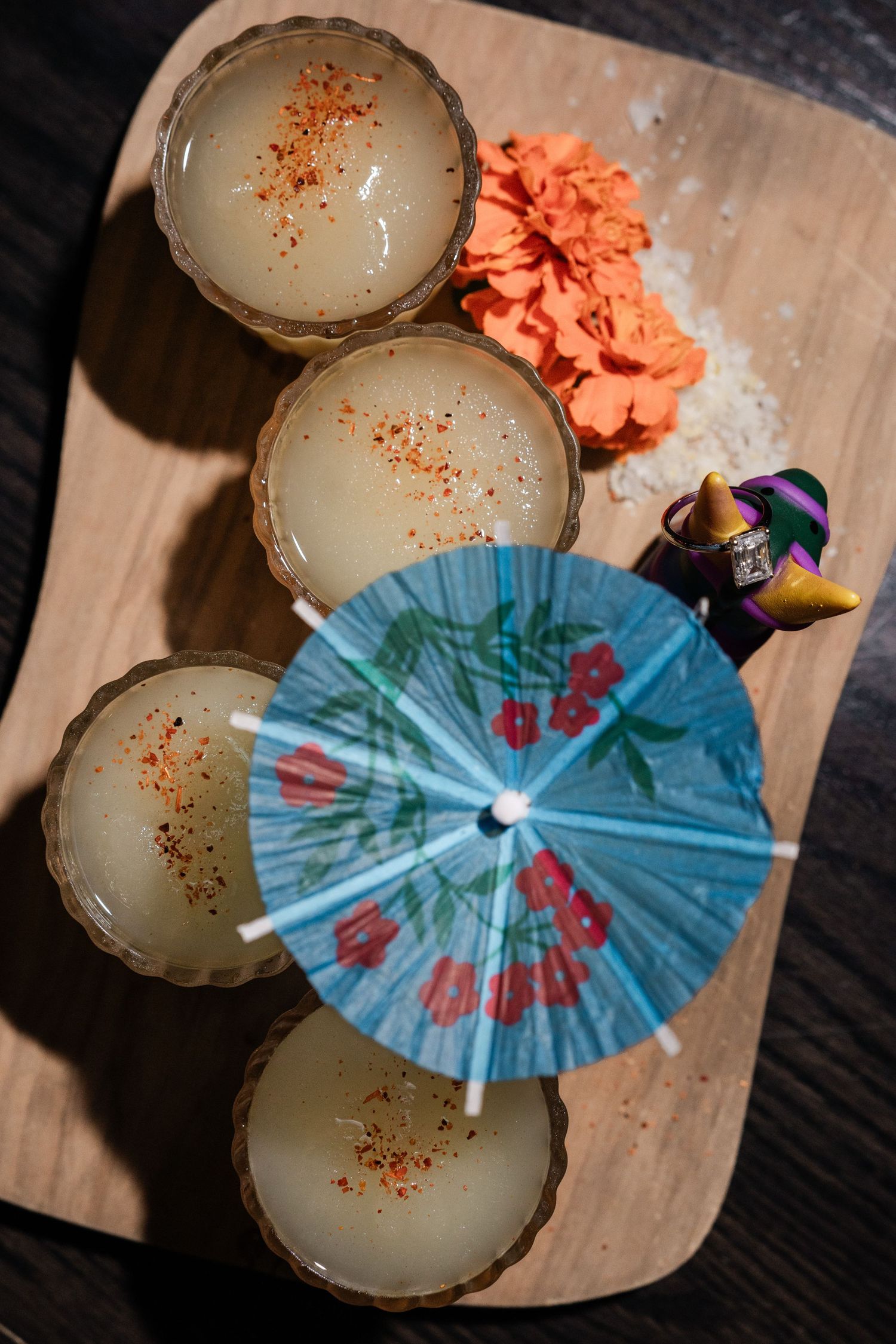 Overhead view of tiki cocktails with chili powder, a paper umbrella, and orange flower on wood.