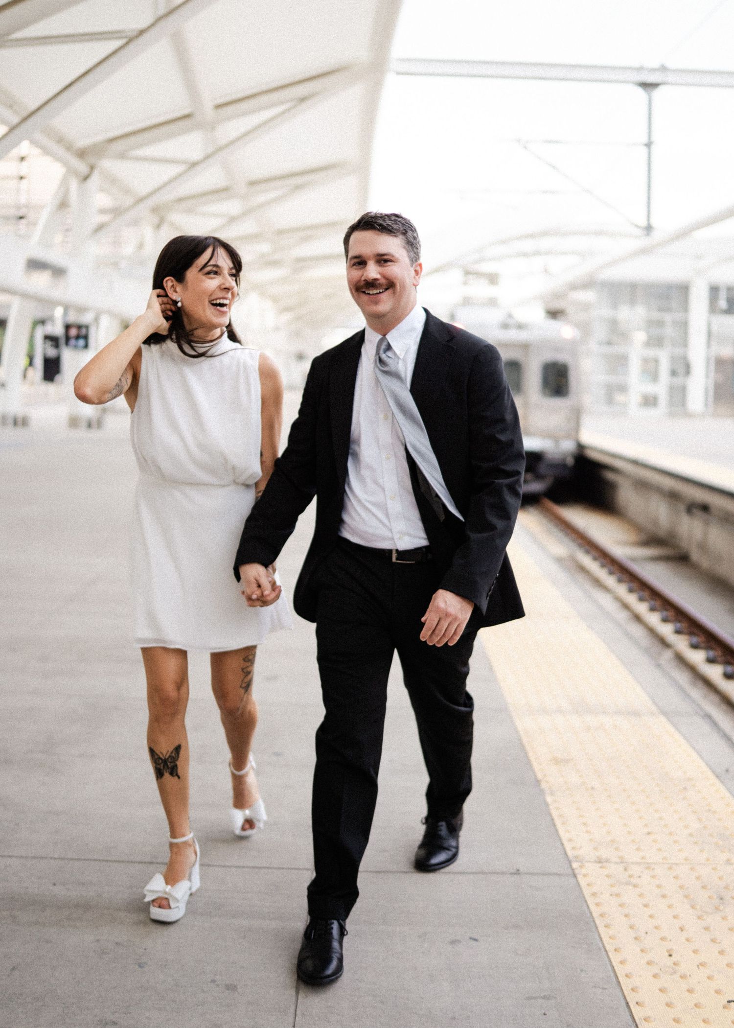 Smiling couple in wedding attire walking hand in hand along a train station platform.