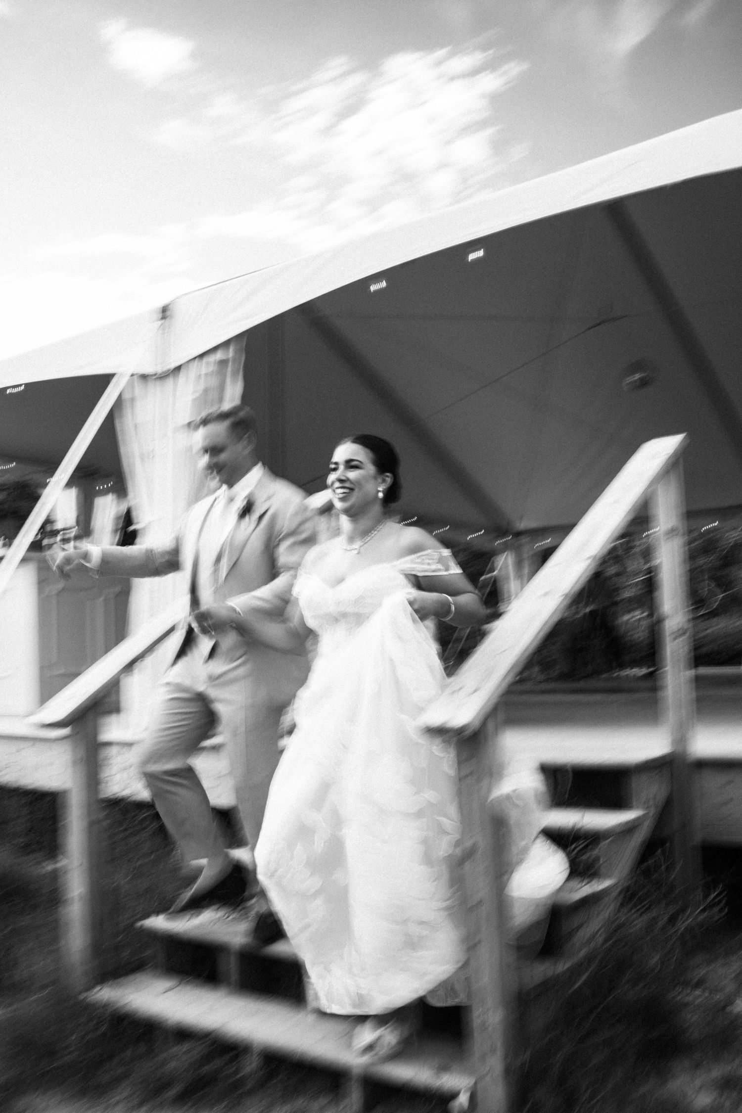 Black and white photo of a joyful bride and groom rushing down wooden steps outside a wedding tent.